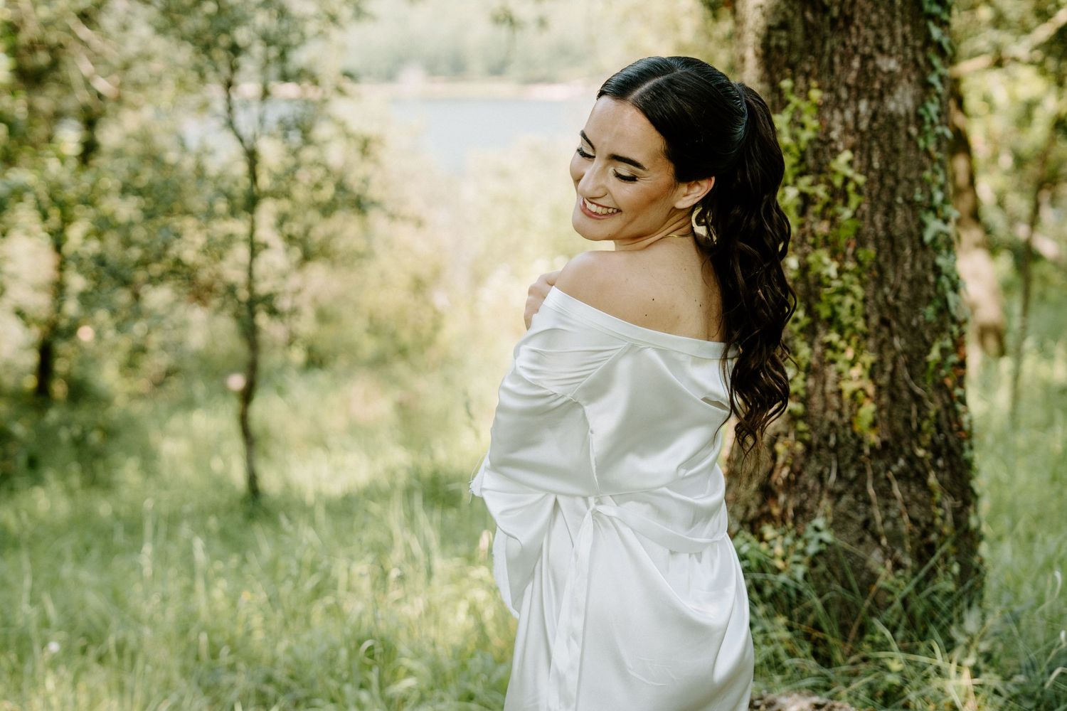 Beautiful bride in a white off-shoulder gown smiling softly while leaning against a tree outdoors.