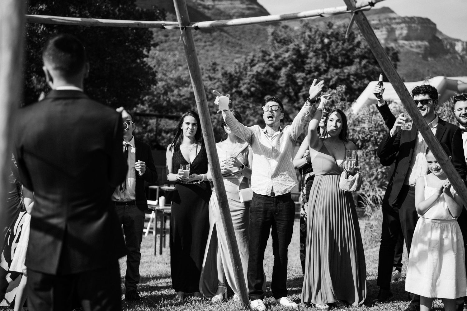 Black and white photo of joyful wedding guests cheering with raised arms during outdoor ceremony.