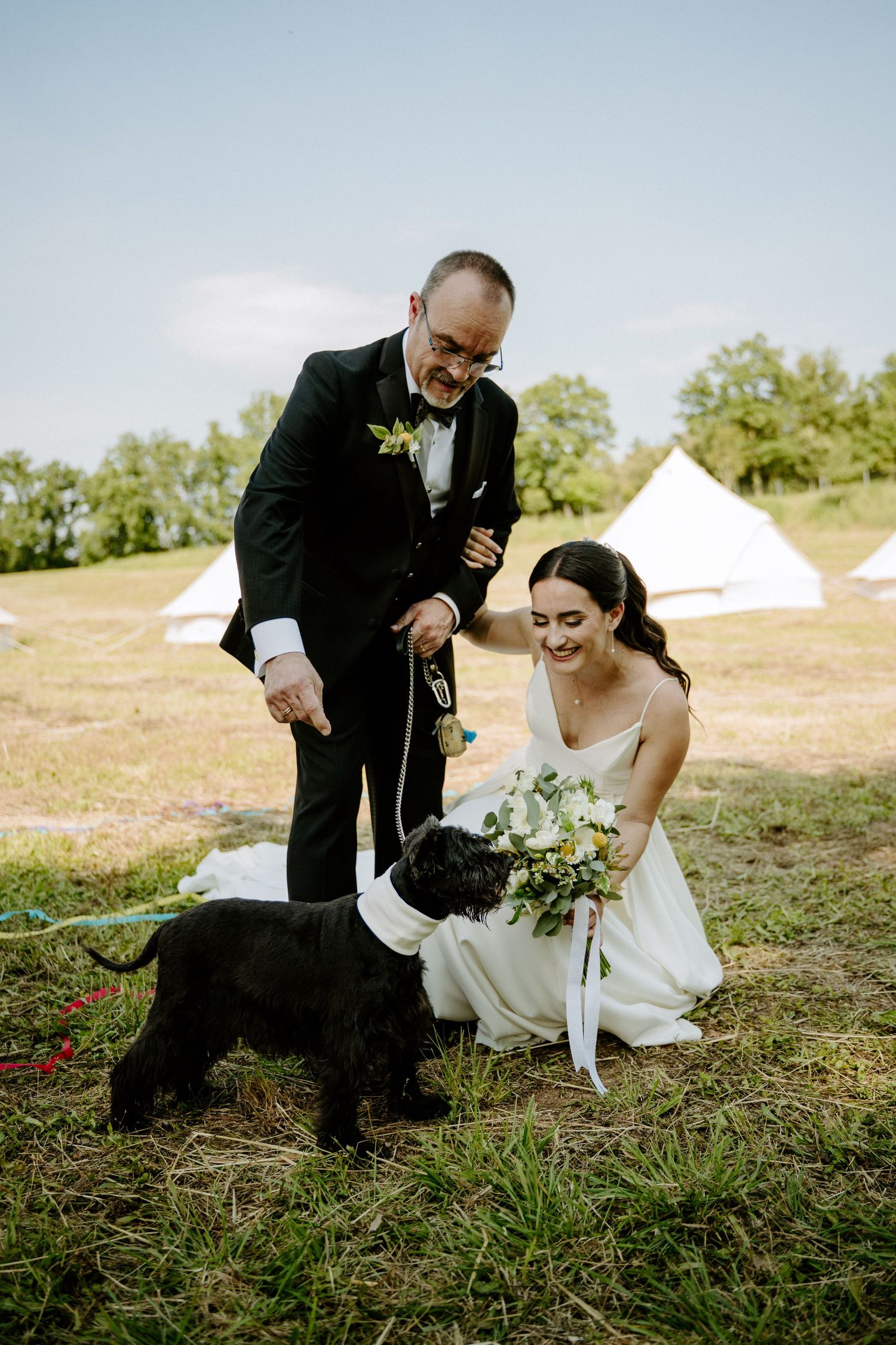 Smiling bride and groom in black tuxedo petting black dog at outdoor wedding venue with white tents.