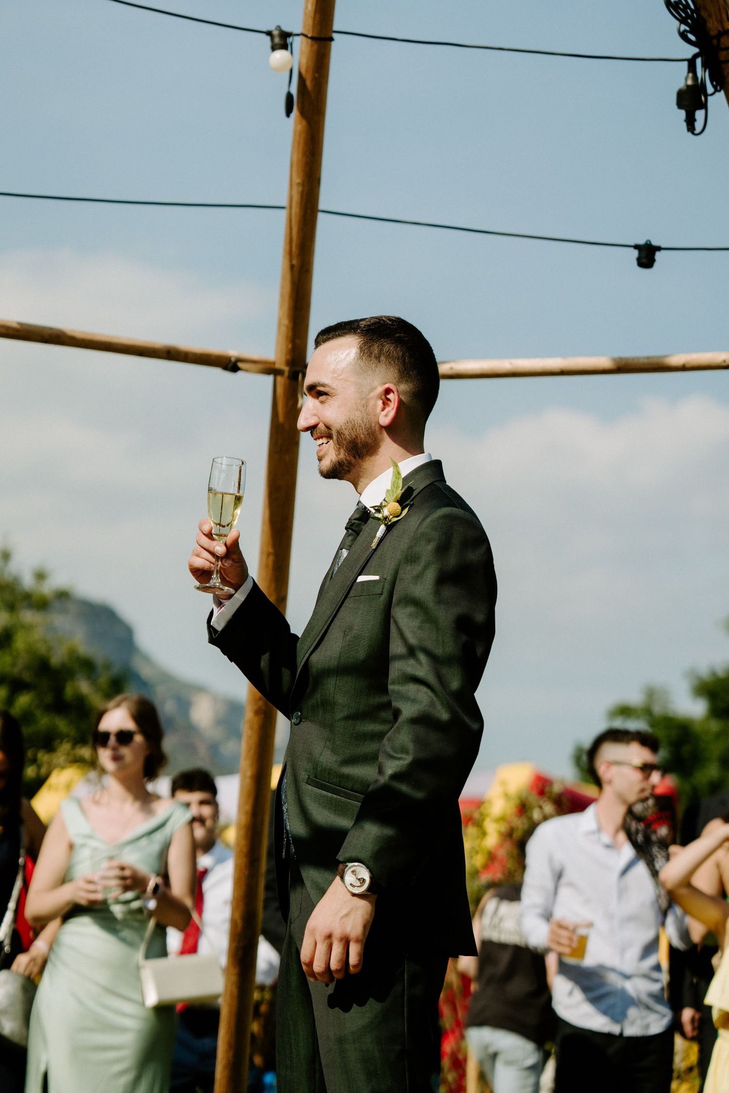Smiling groom in dark suit raising champagne glass toast at outdoor wedding celebration.