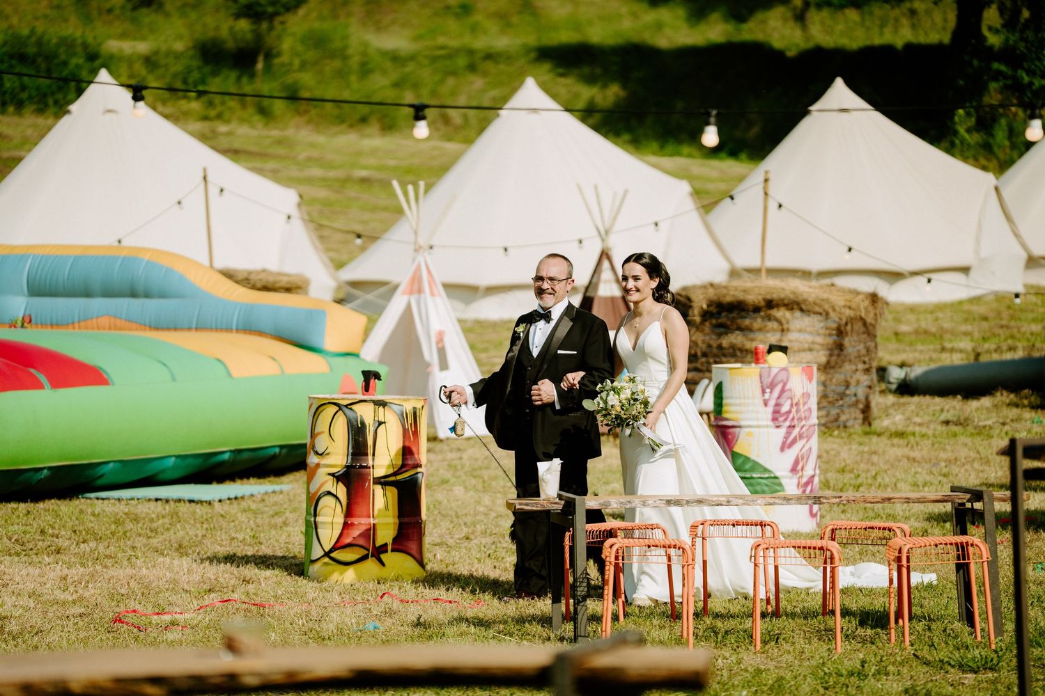 Bride and groom pose together in field surrounded by glamping tents.