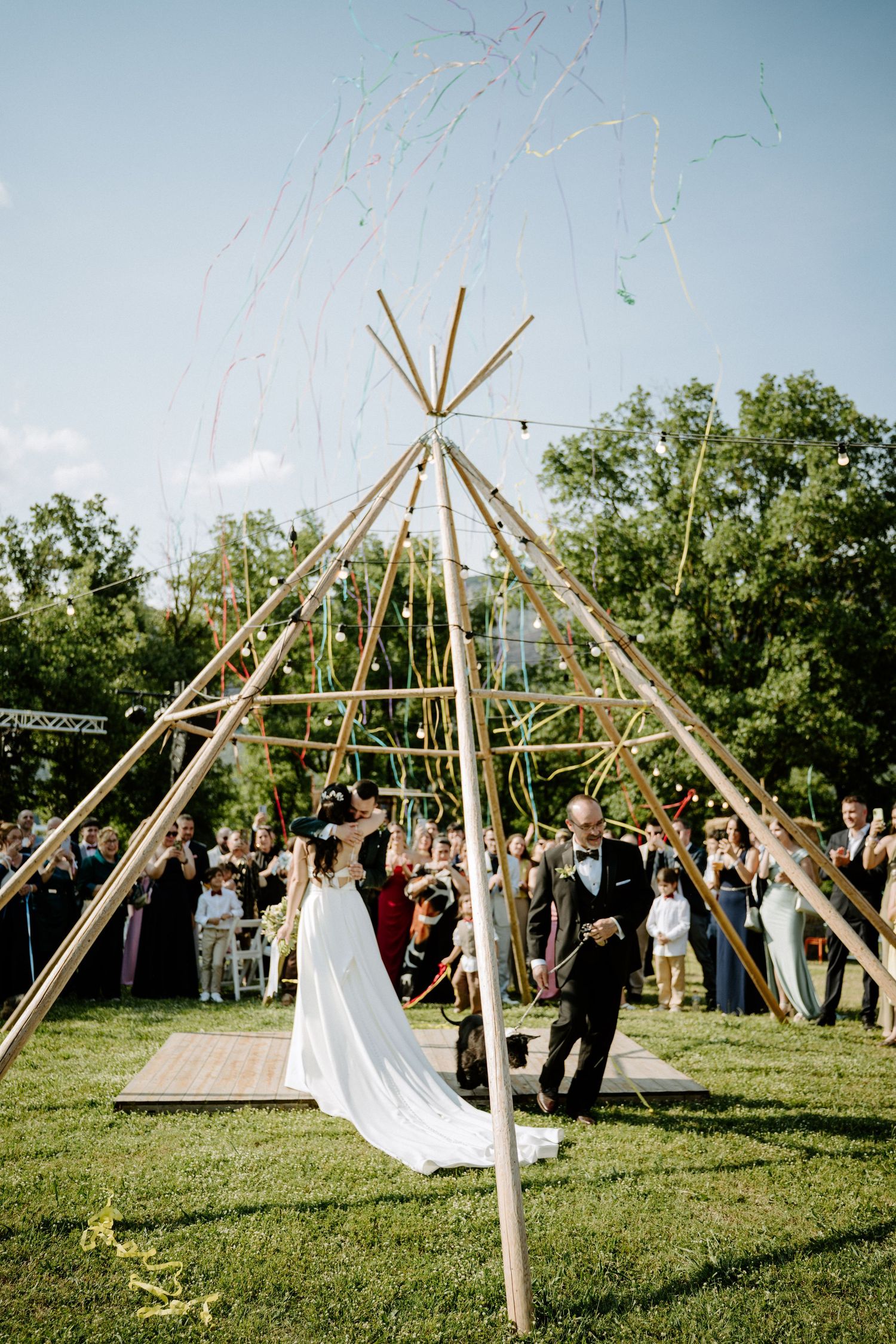 A bride and groom exchange vows under a tall bamboo teepee structure at an outdoor wedding ceremony.