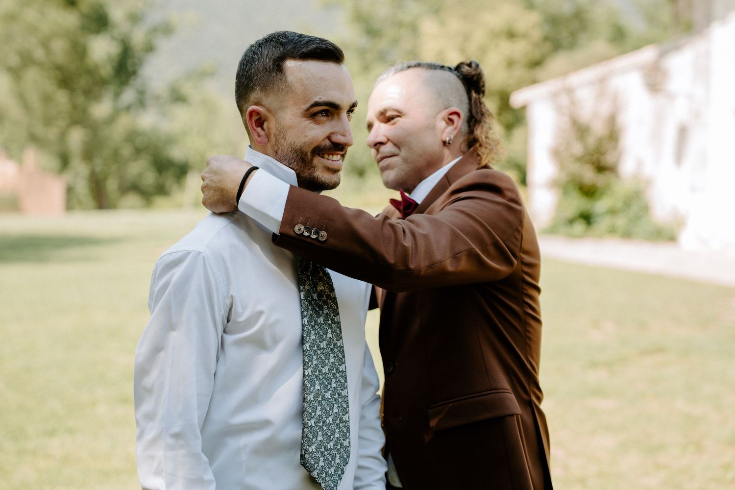 A man in a brown suit adjusts the tie of a smiling groom in a light blue shirt outdoors.