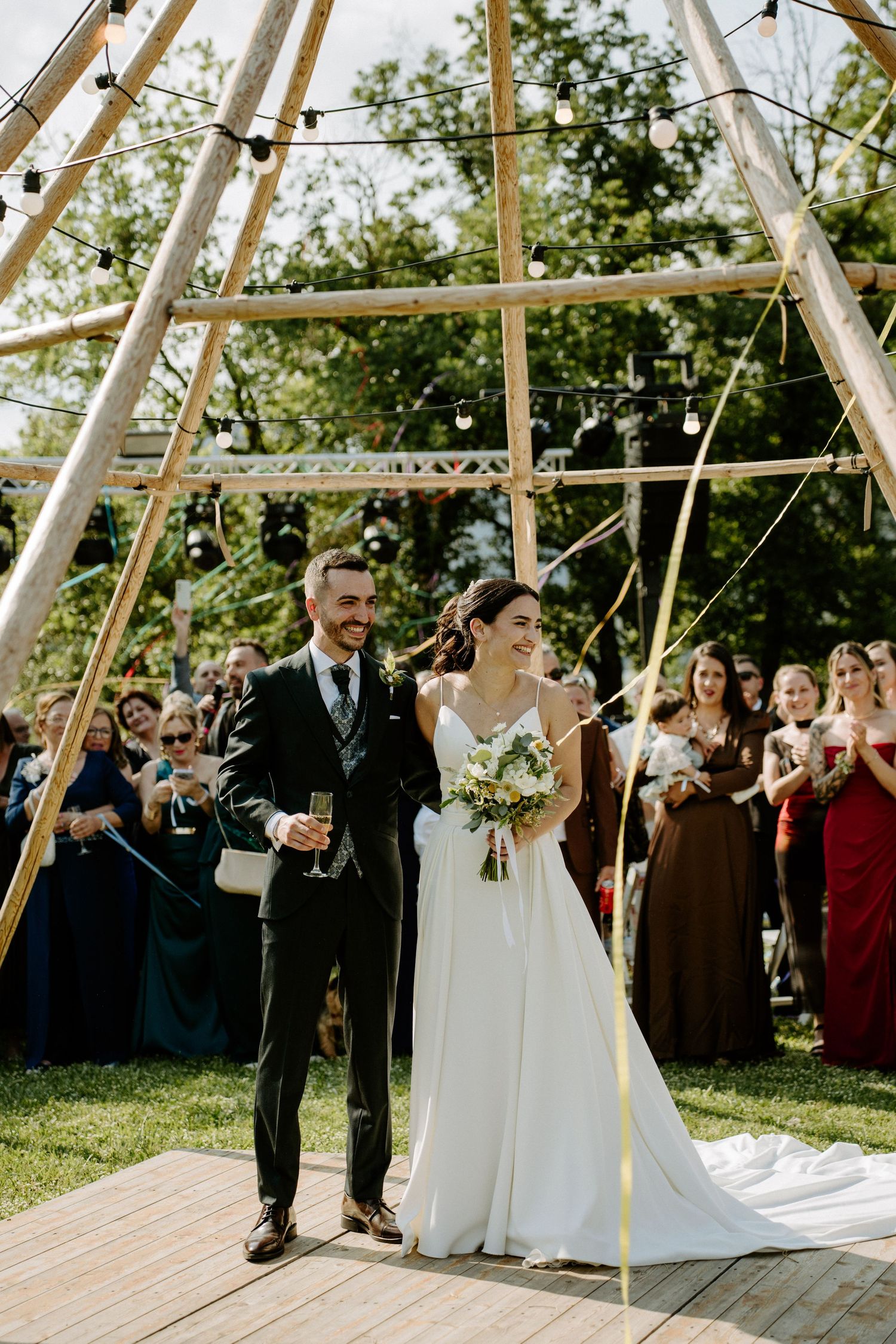 Newlyweds walking down the aisle under a geometric wooden arch surrounded by cheering wedding guests.