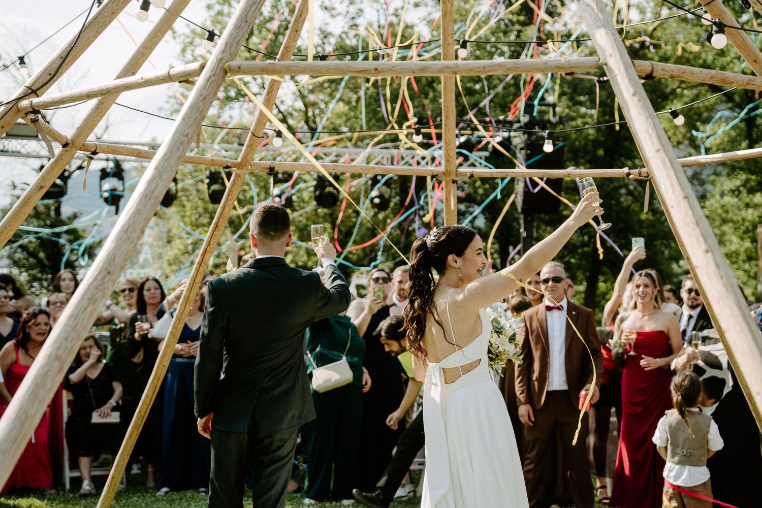 Bride and groom dancing under geometric wooden arch with guests celebrating at outdoor wedding.