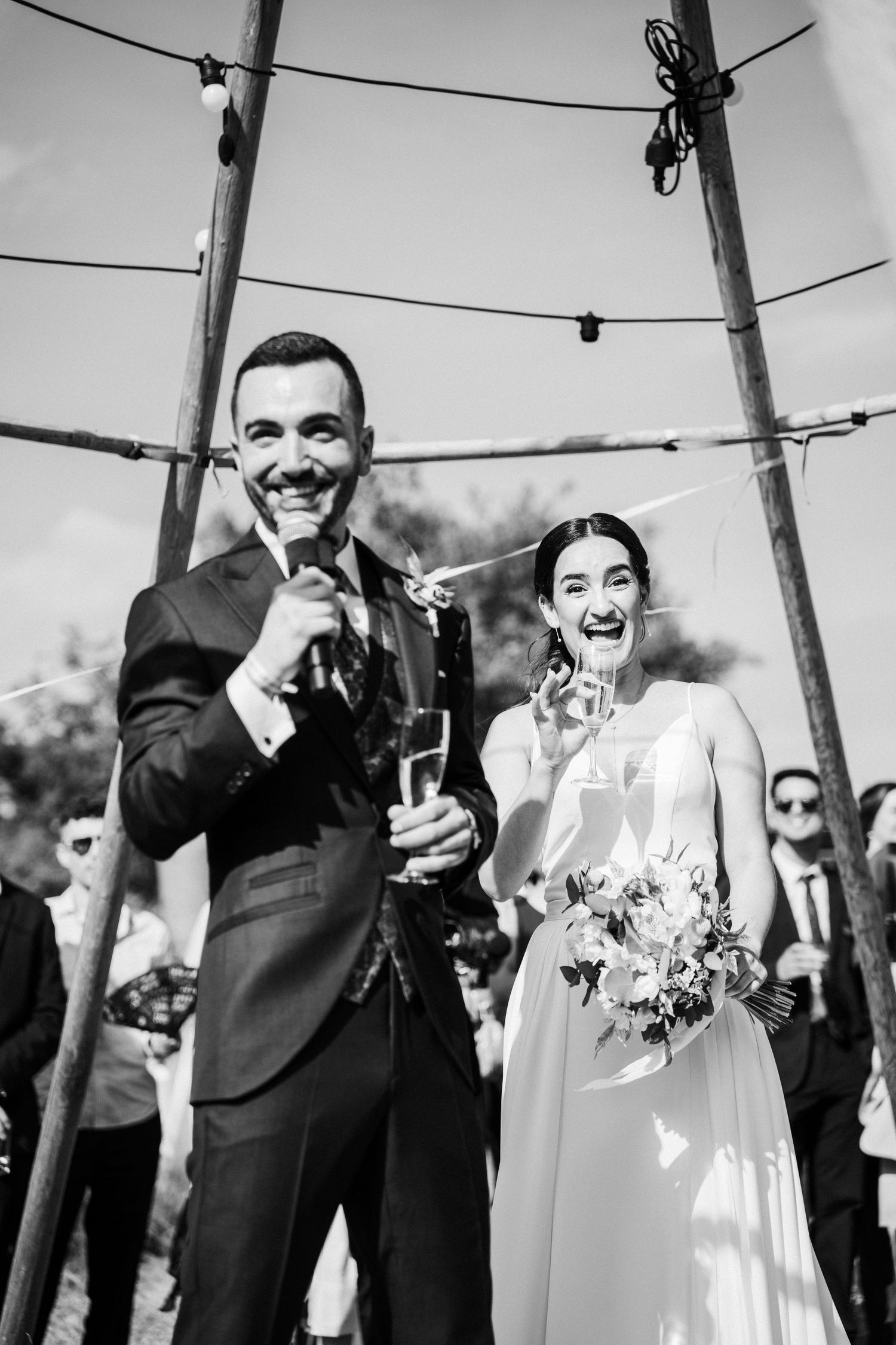 Joyful bride and groom laughing and celebrating with champagne at outdoor wedding ceremony in black and white photo.