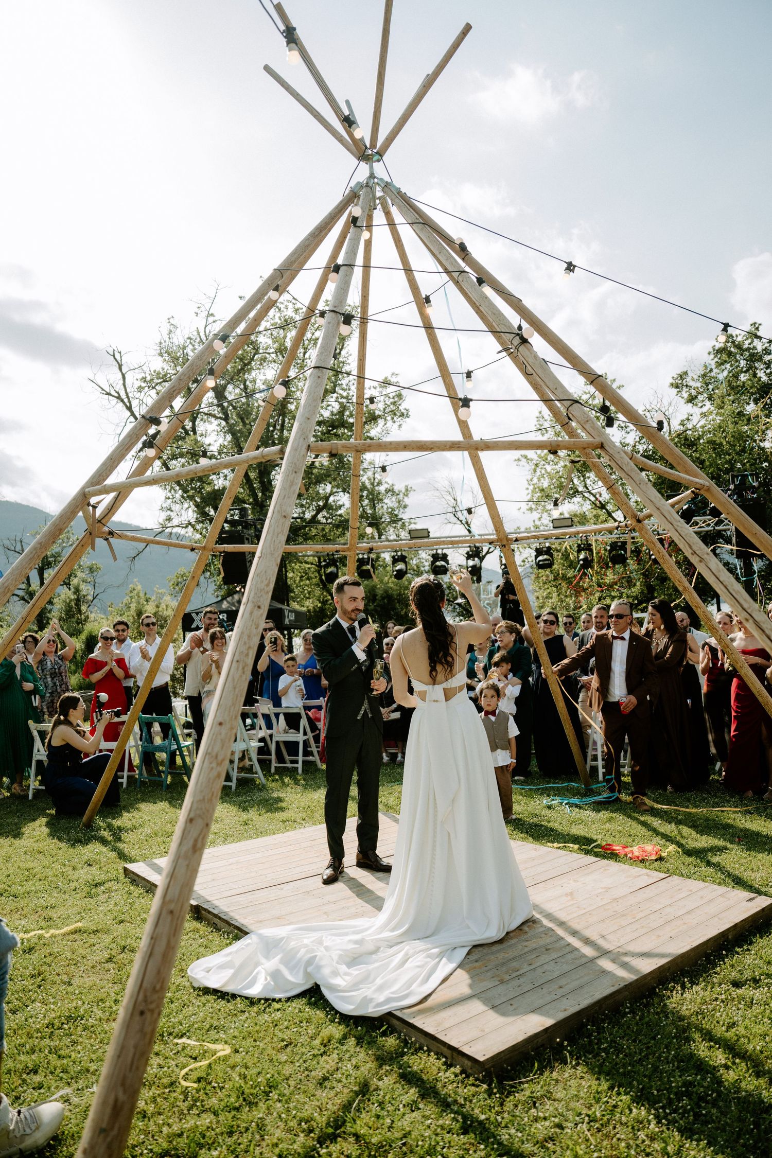 Bride and groom exchanging vows under rustic wooden tipi structure at outdoor garden wedding ceremony.