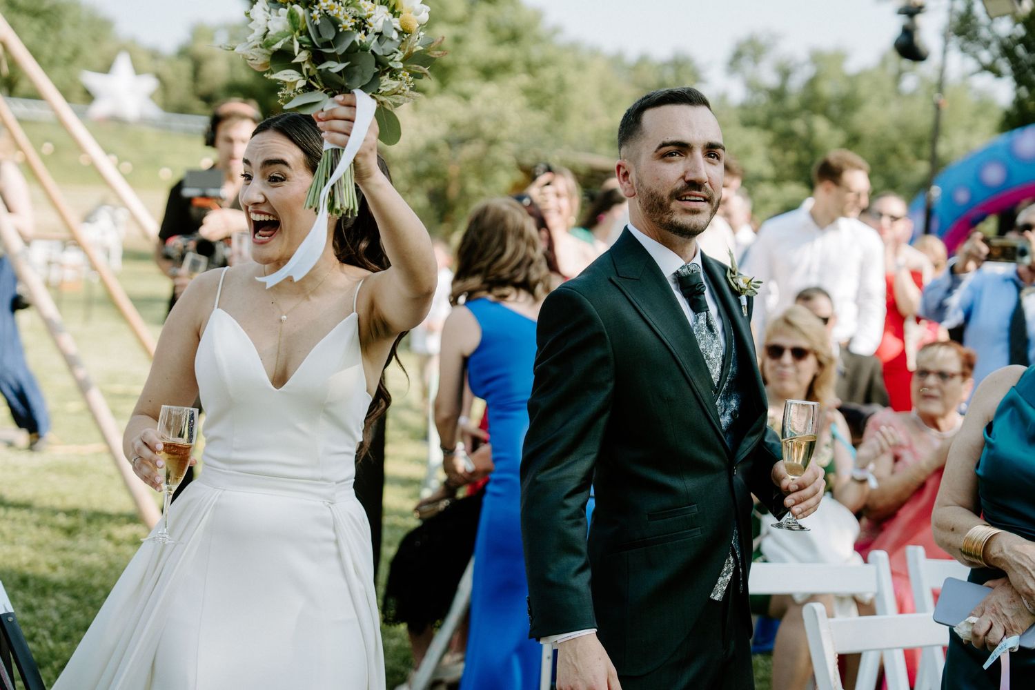 Joyful bride raising bouquet with groom holding champagne after outdoor wedding ceremony.