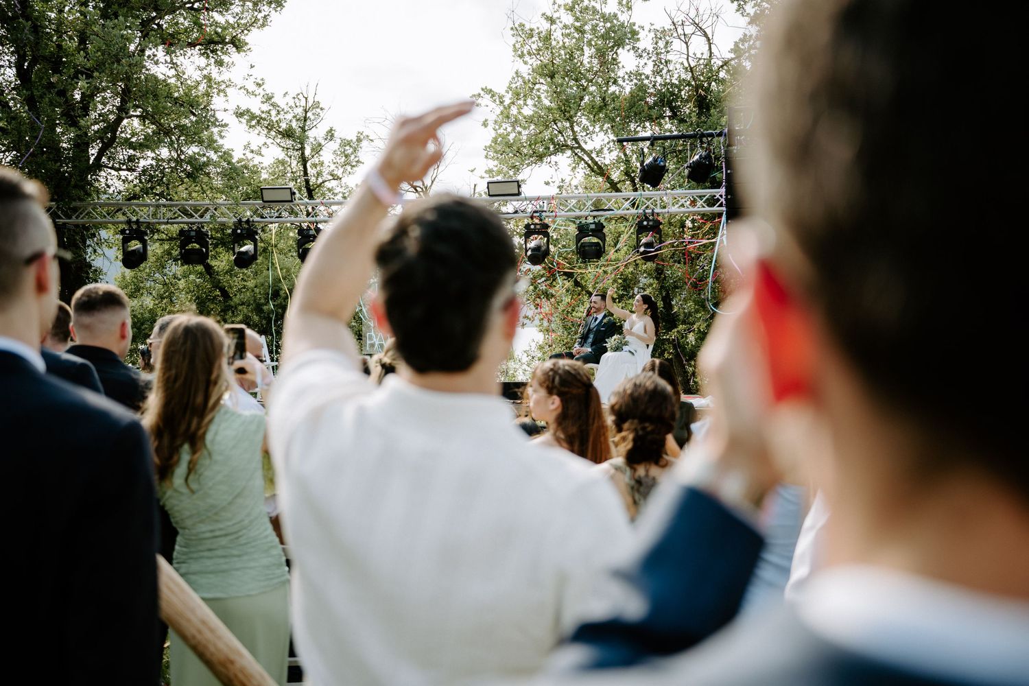 Guests celebrating at an outdoor wedding ceremony with a stage and greenery in the background.