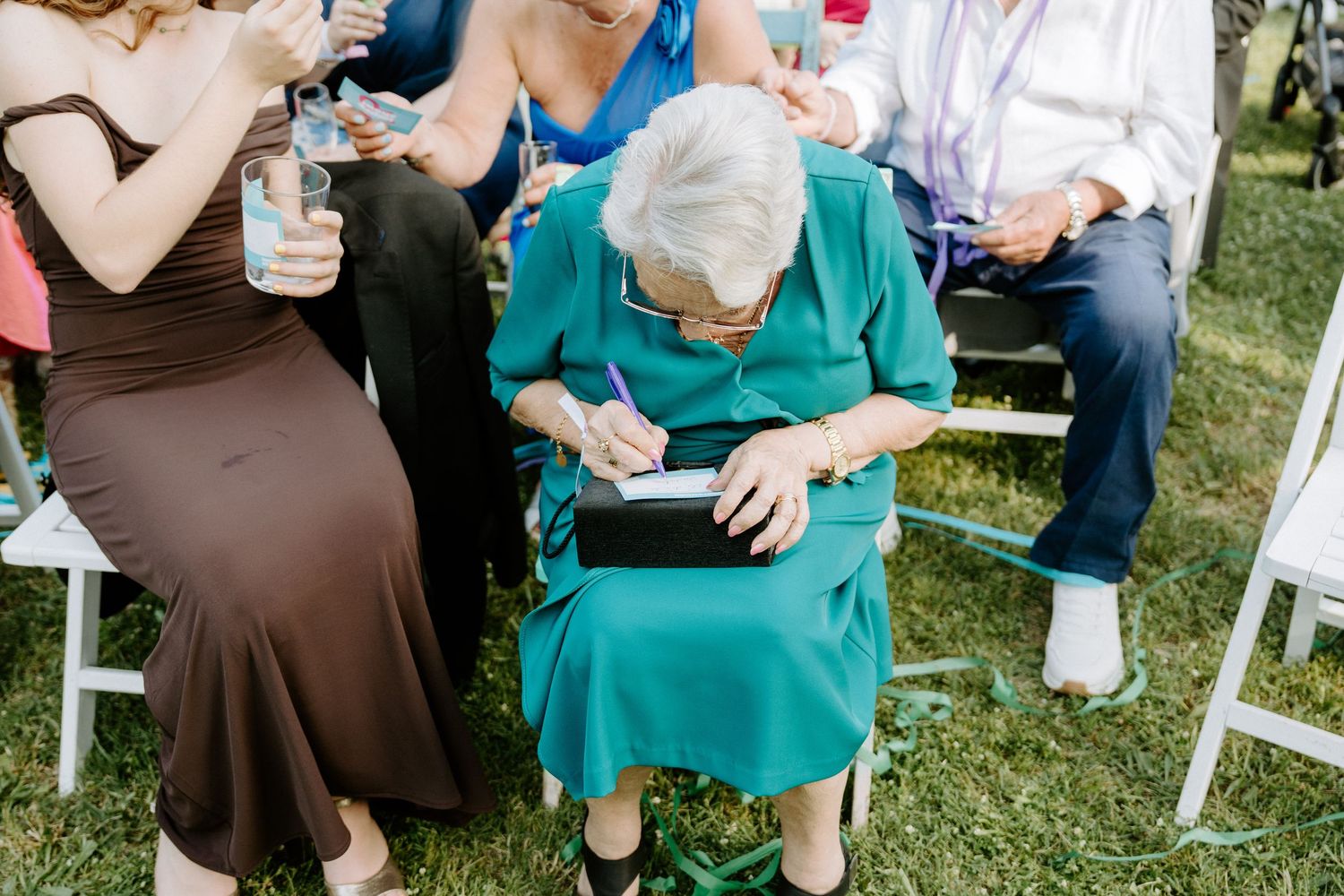 Elderly woman in teal dress writing in notebook while seated among wedding guests at outdoor ceremony.