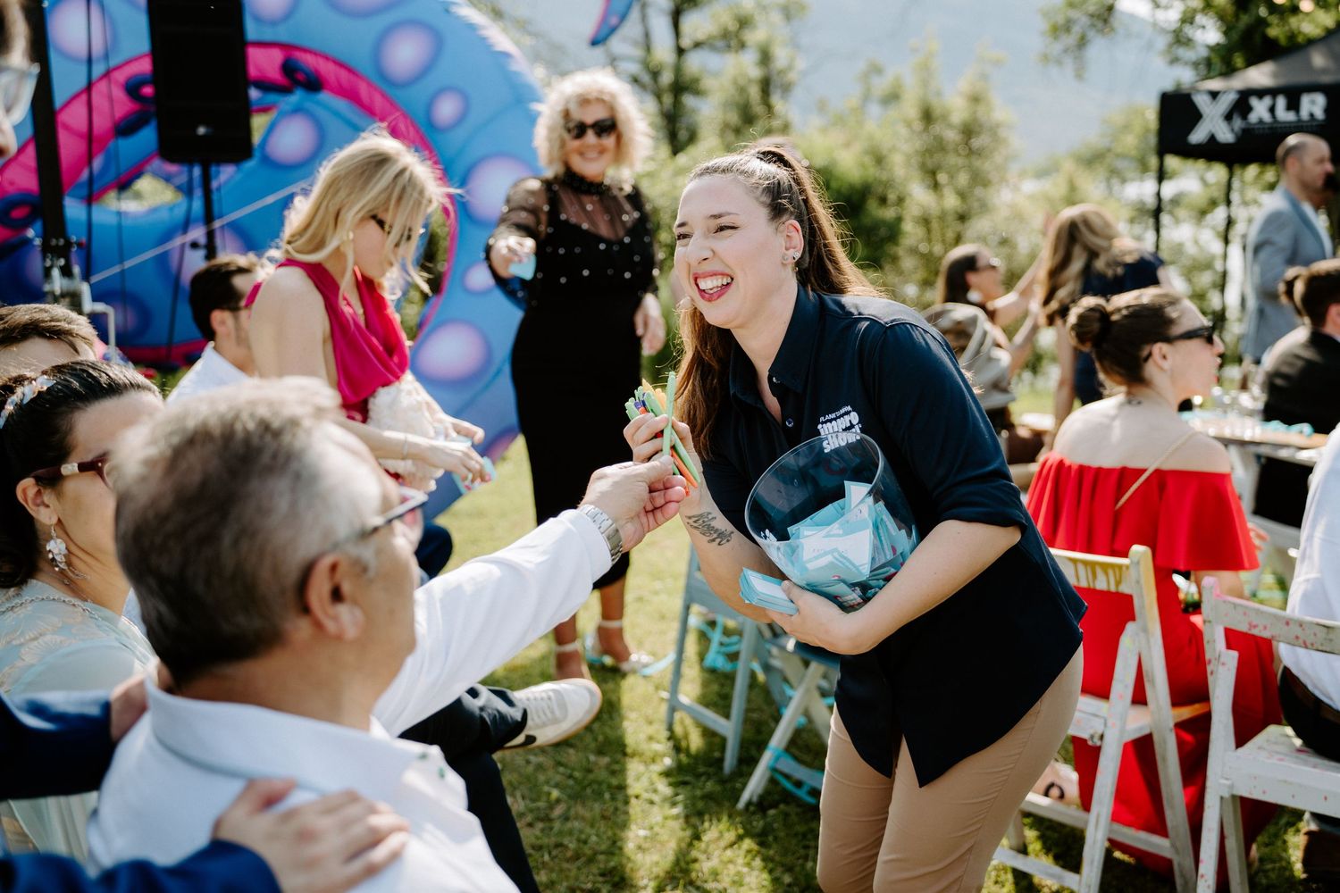 Smiling wedding staff member in black shirt handing items to seated guests at outdoor reception.