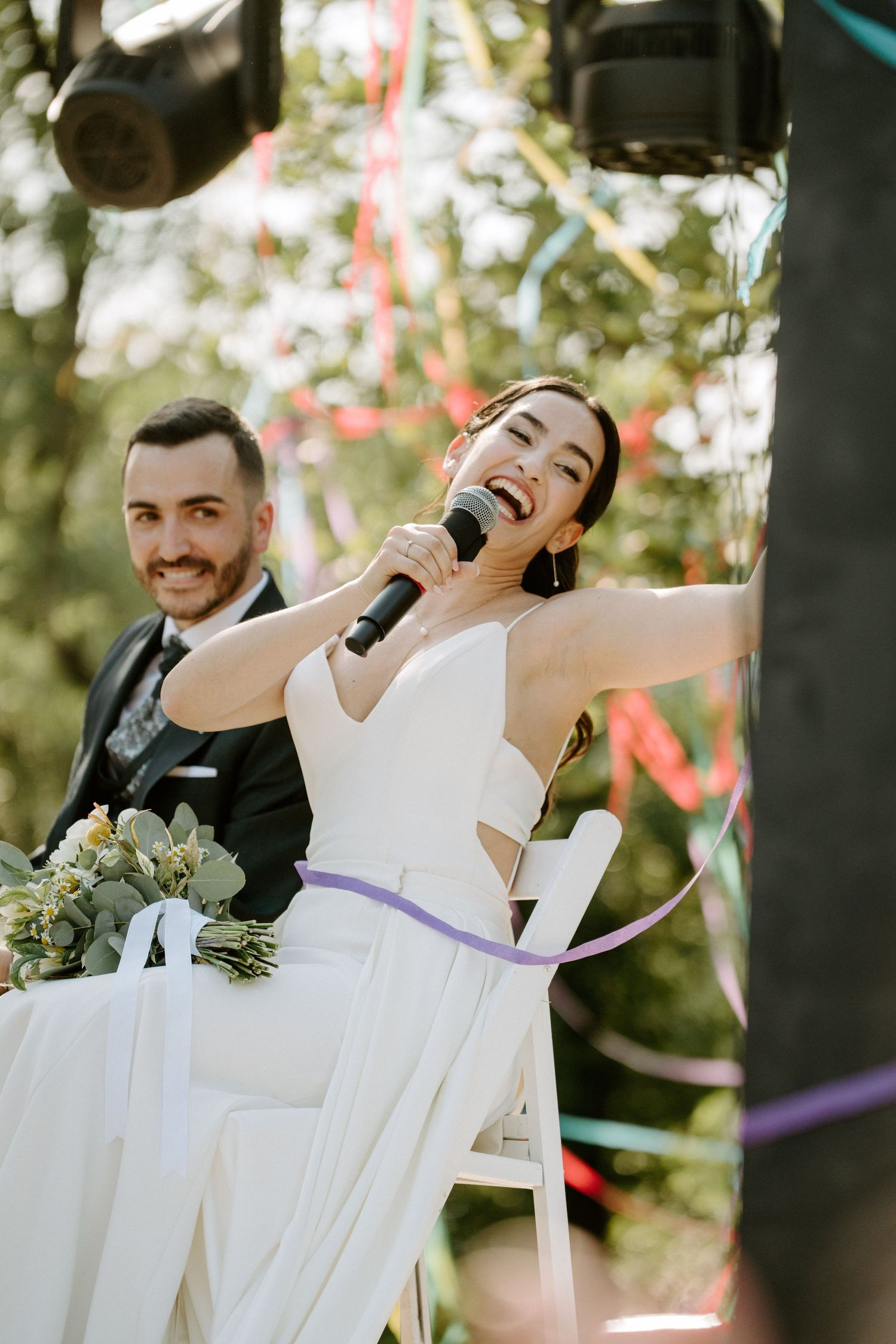 Radiant bride singing into microphone at outdoor wedding reception while groom smiles beside her.