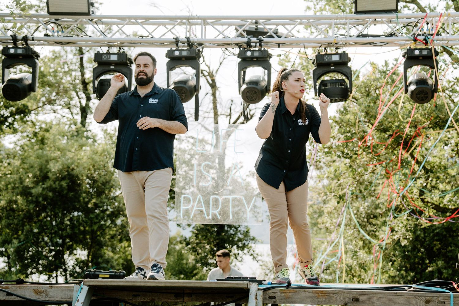 Two wedding MCs in black shirts performing on a decorated outdoor stage at a party.