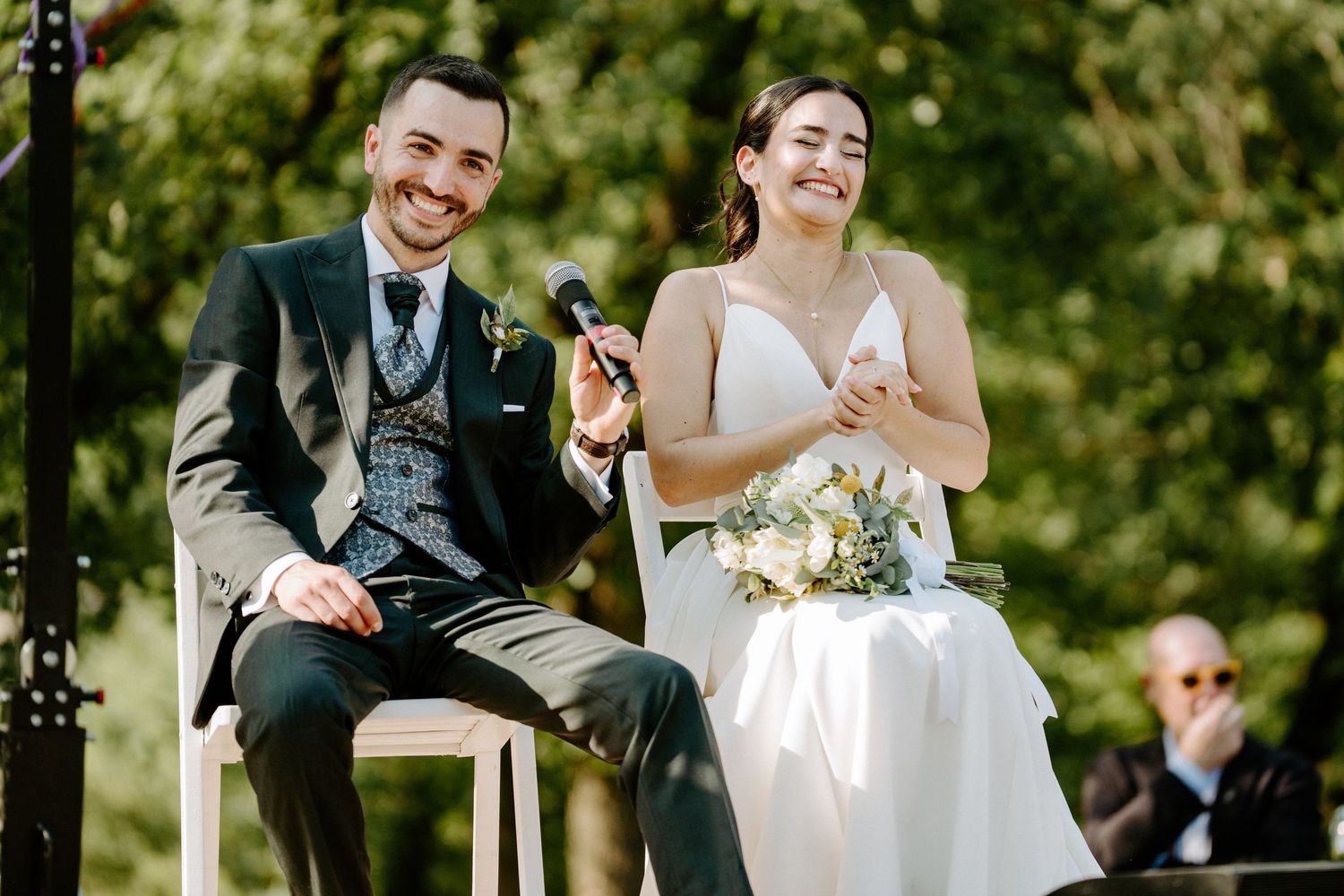 Smiling bride and groom sitting together during outdoor wedding ceremony, groom holding microphone.