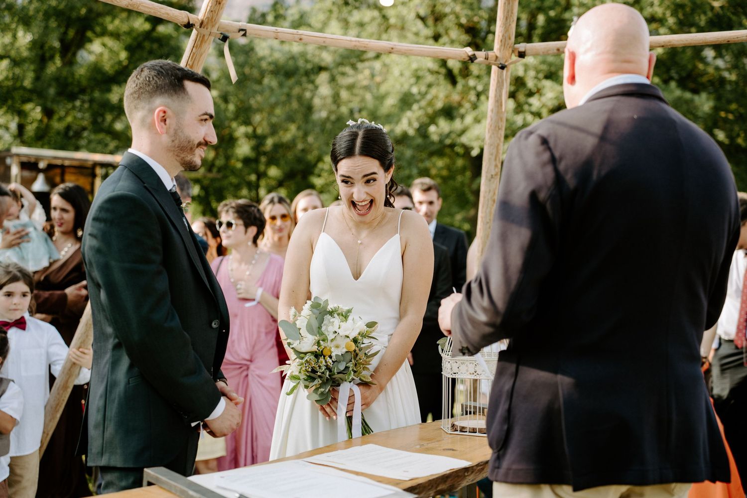 Bride holding greenery bouquet laughing with groom during outdoor woodland wedding ceremony.
