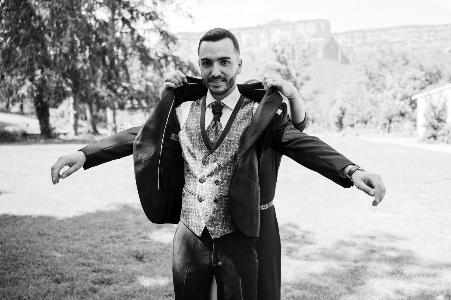 Dapper groom in black suit and patterned vest poses with arms outstretched in black and white photo.
