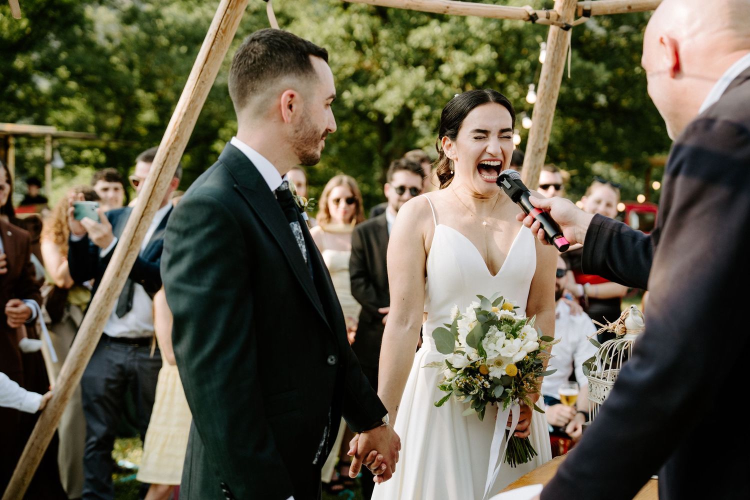 Laughing bride in white gown holds bouquet with groom during joyful outdoor wedding ceremony.