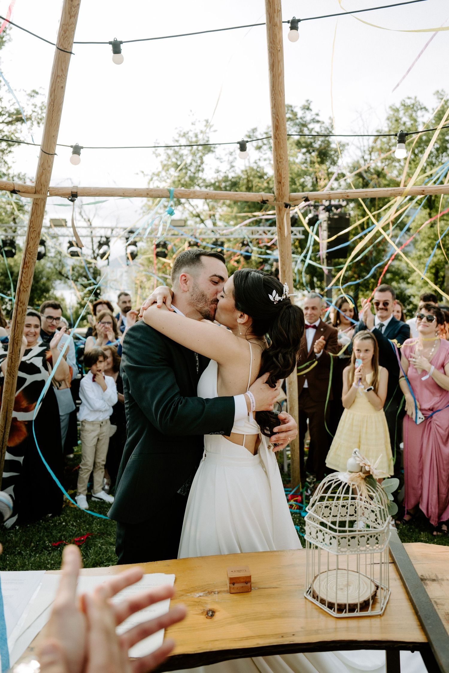 Bride and groom sharing first kiss under colorful streamers at outdoor tipi wedding ceremony with cheering guests.