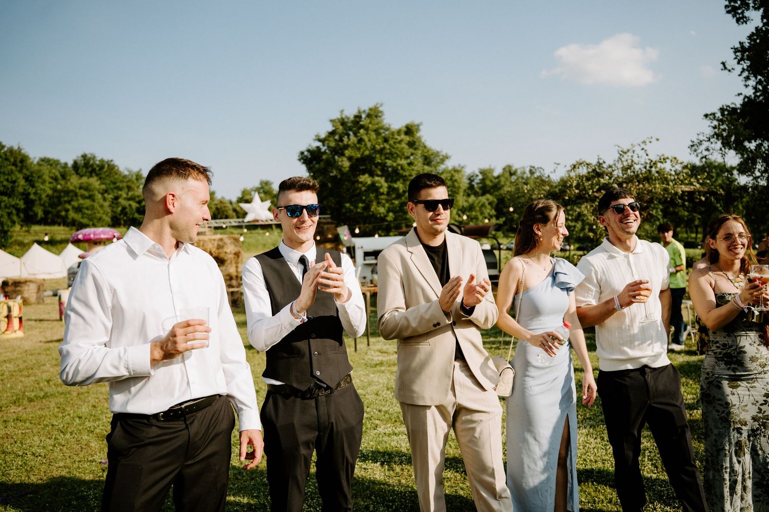 Groomsmen in suits and sunglasses celebrating outdoors at sunny wedding reception.