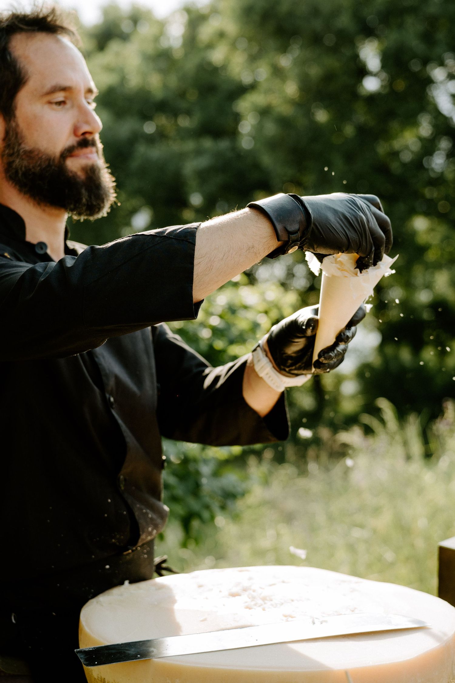 Bearded chef in black gloves grating fresh cheese over wheel outdoors in sunlight.
