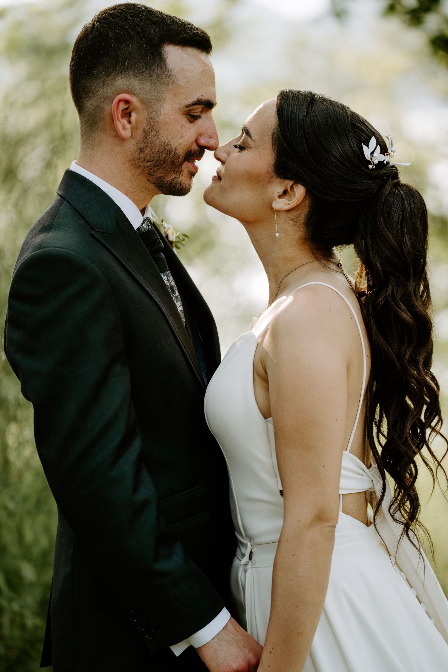 Bride and groom sharing an intimate nose-to-nose moment while holding hands outdoors on wedding day.