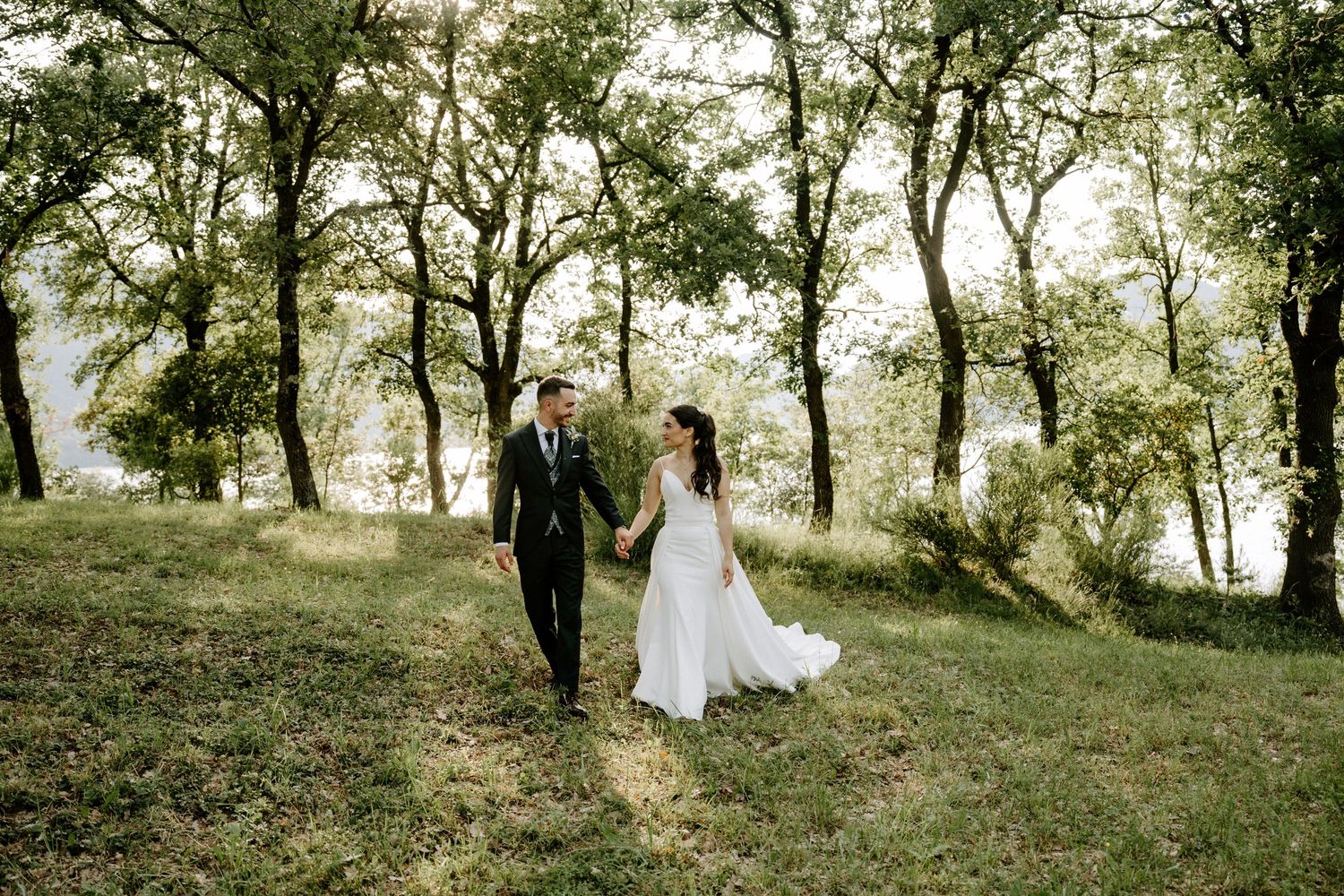 Bride and groom walk hand-in-hand through a lush green forest clearing on their wedding day.