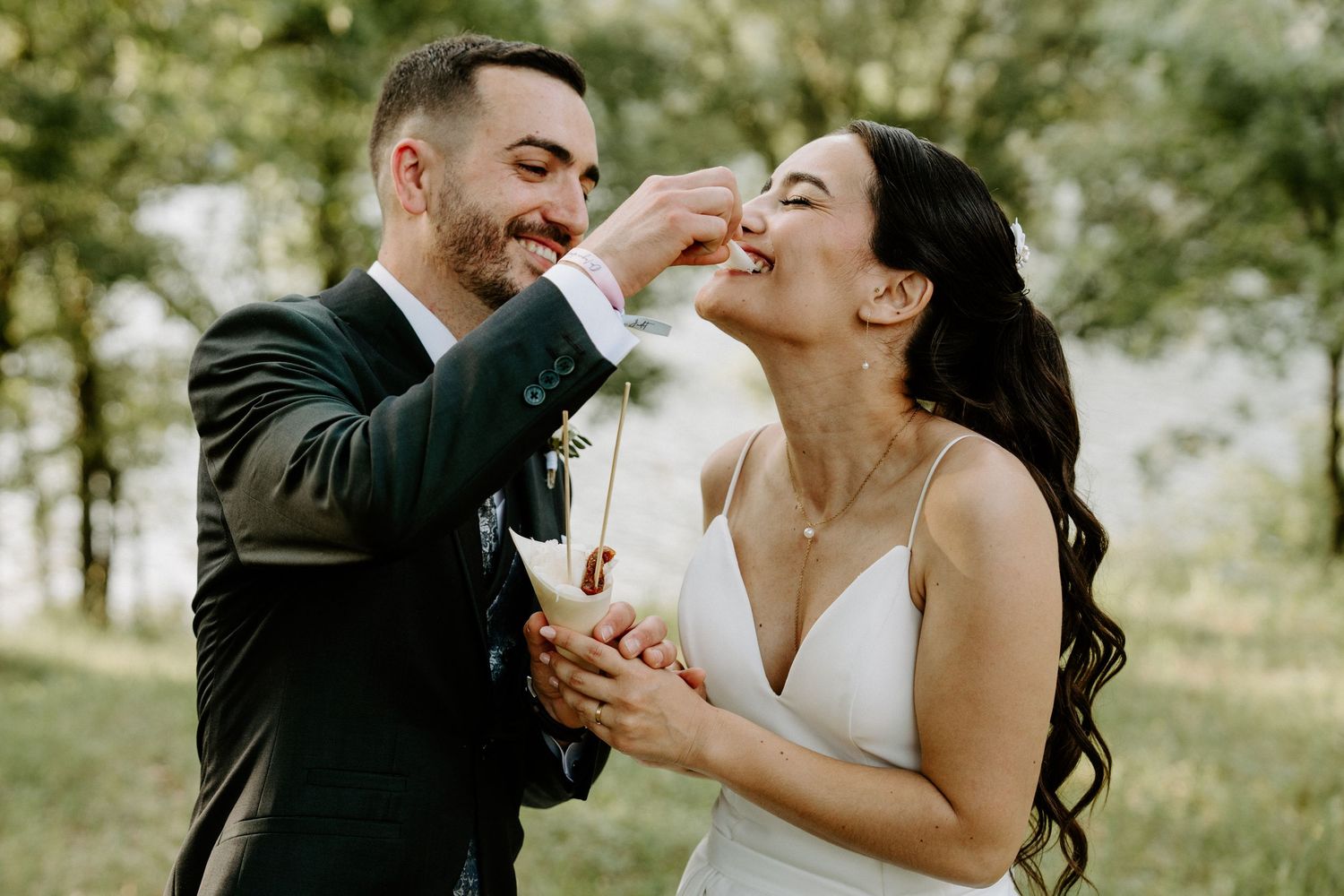 Groom playfully feeding smiling bride at outdoor wedding portrait session in green park.