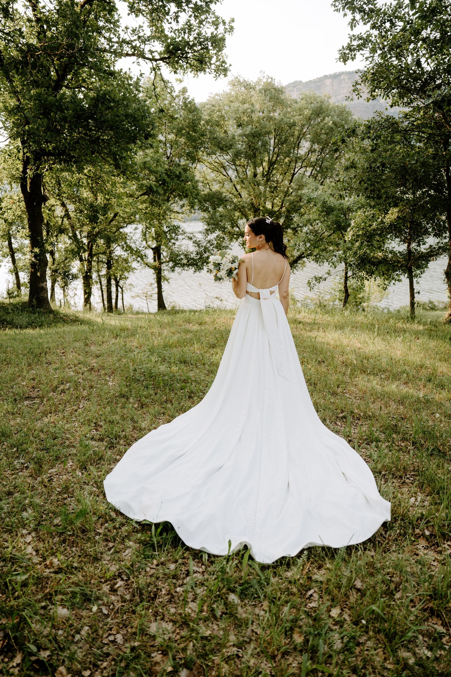 Bride in elegant white gown with long train standing in lush green outdoor setting near lake.