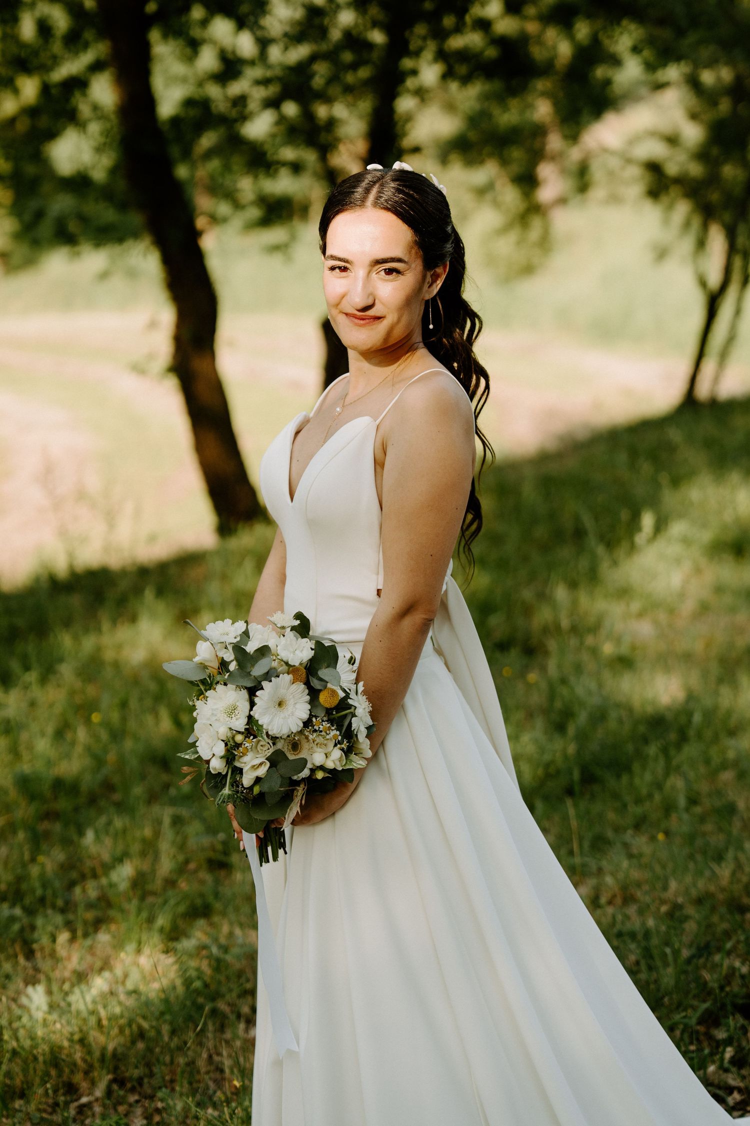 Bride in white gown holding bouquet of white flowers, smiling outdoors in green garden setting.
