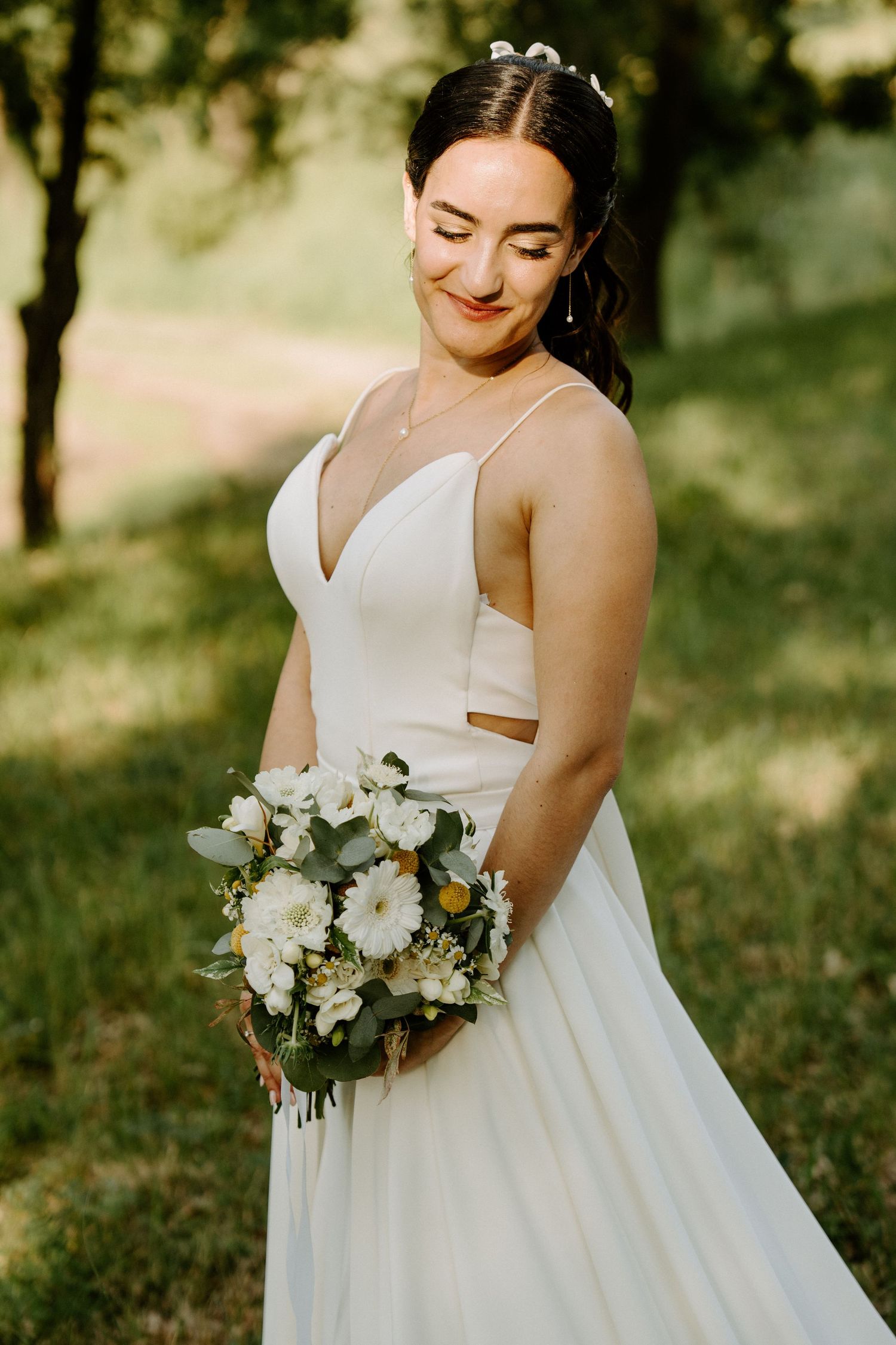 A smiling bride in a white spaghetti-strap gown holds a white floral bouquet in a garden setting.