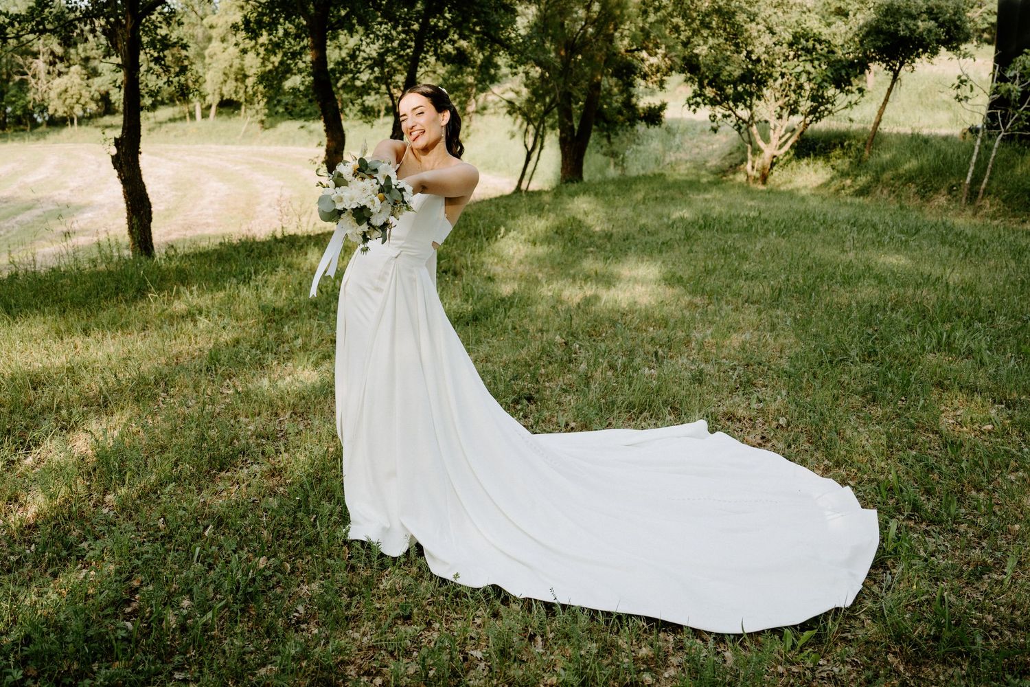 Joyful bride in white gown with long train twirling with bouquet in sunny green meadow.