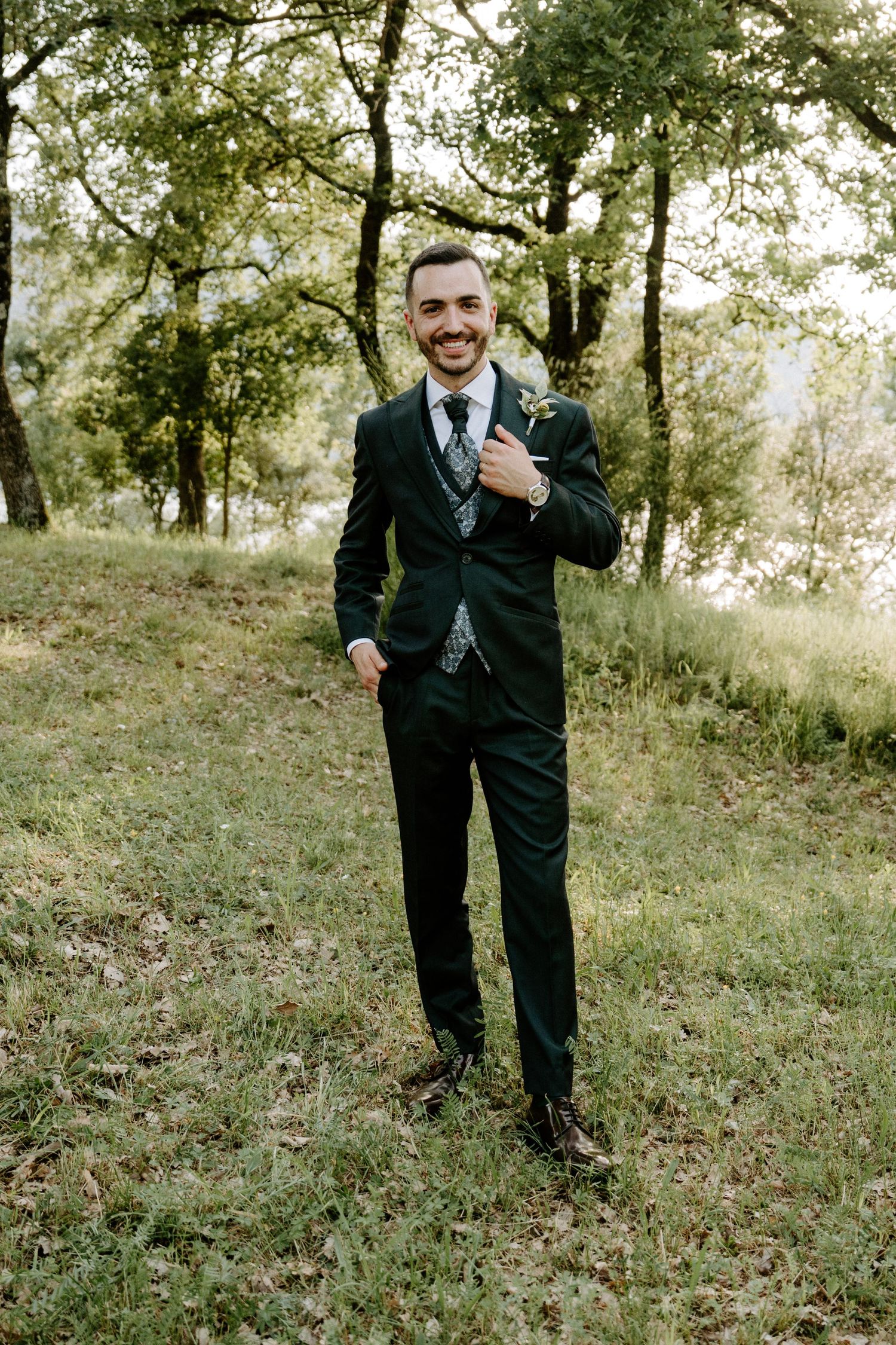 Smiling groom in dark suit with floral tie stands outdoors on grass with trees in background.