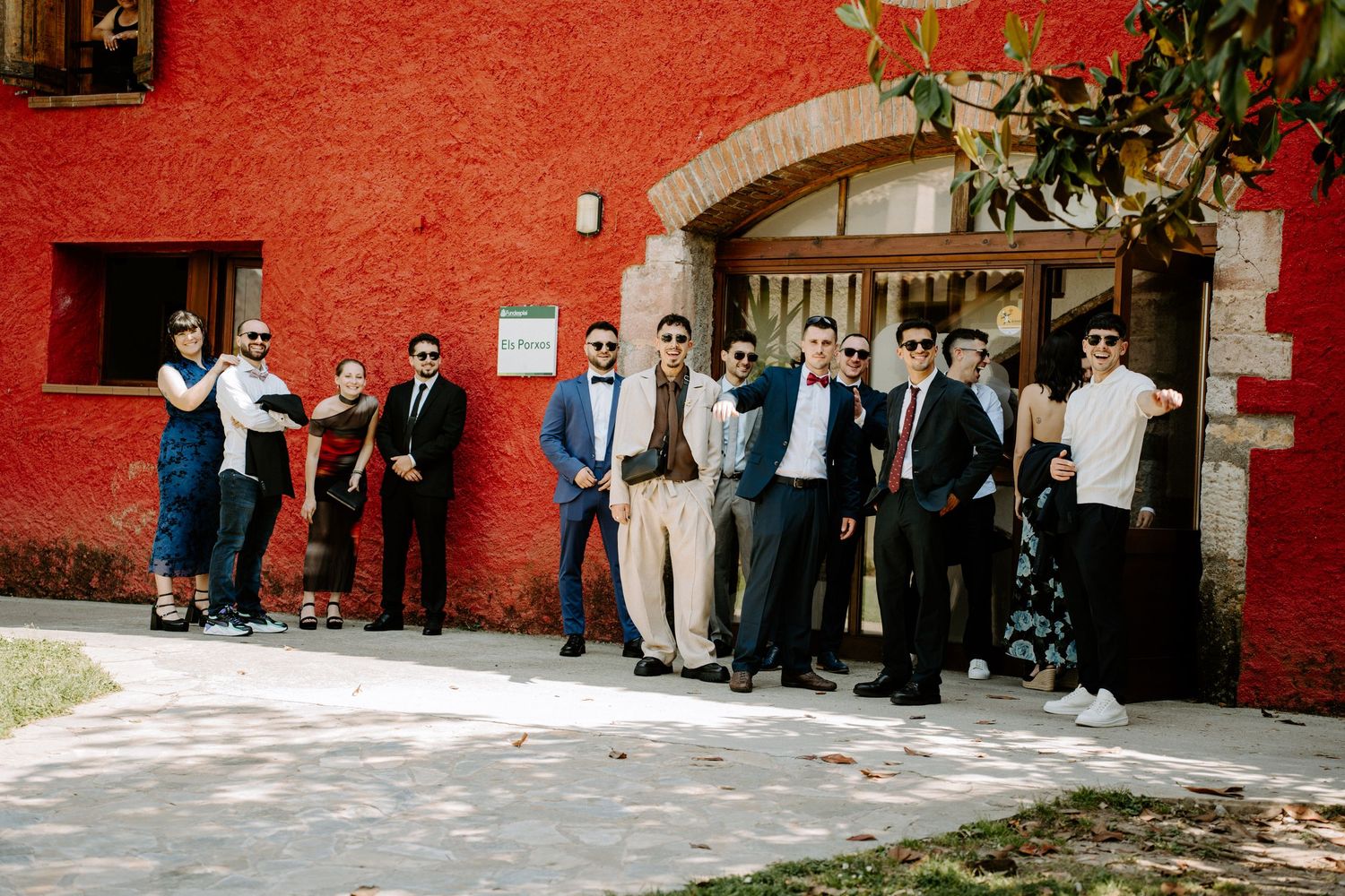 Group of elegantly dressed wedding guests posing outside a rustic red building on a sunny day.