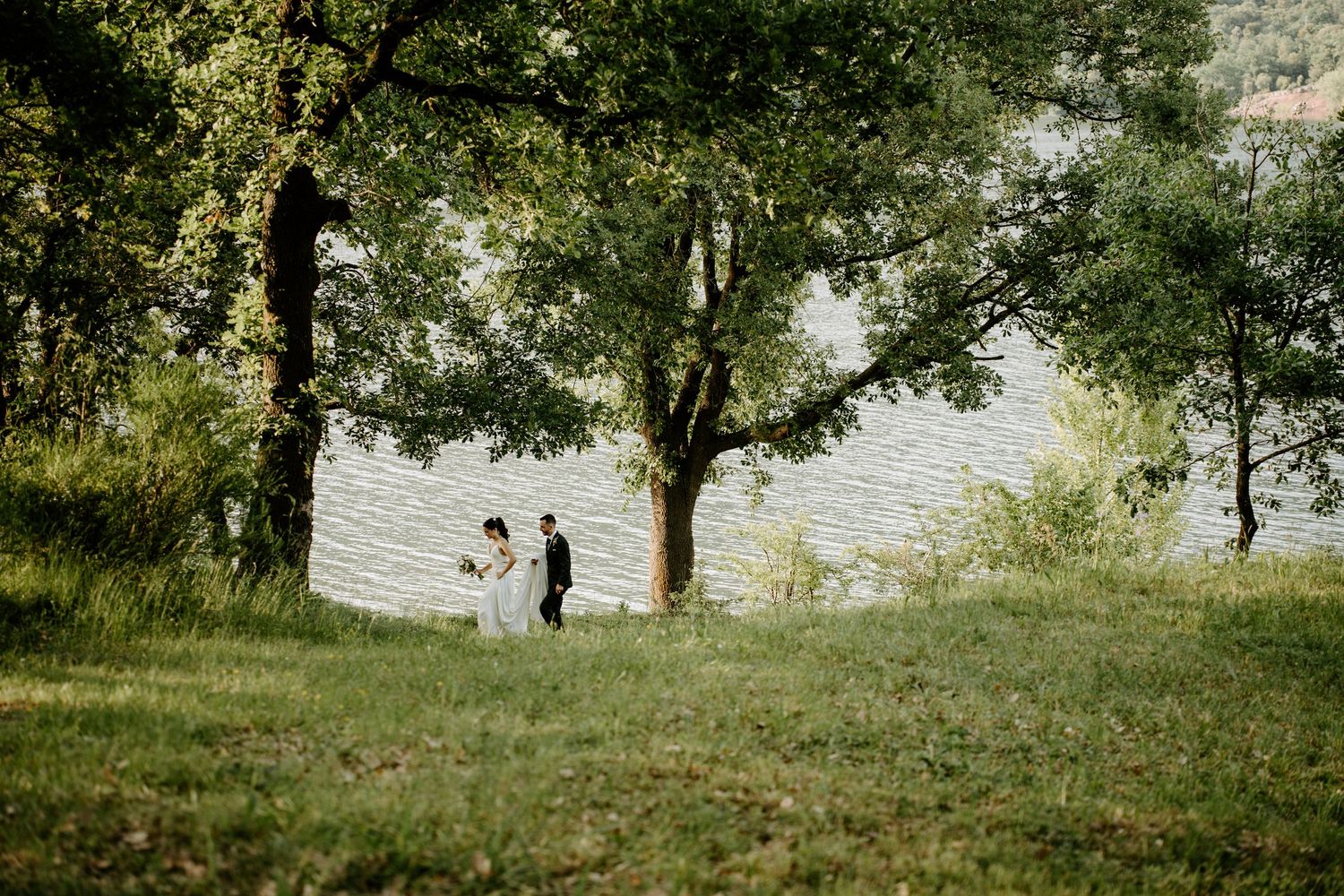 Bride and groom share intimate moment beneath large tree by serene lakeside in nature.