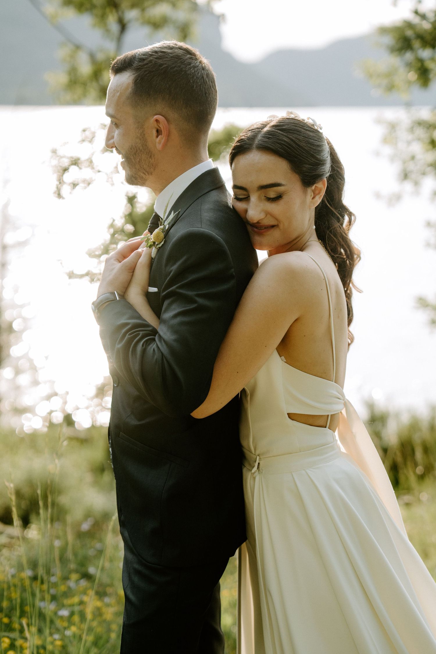 Bride embracing groom from behind near a sunlit lake surrounded by lush green trees.