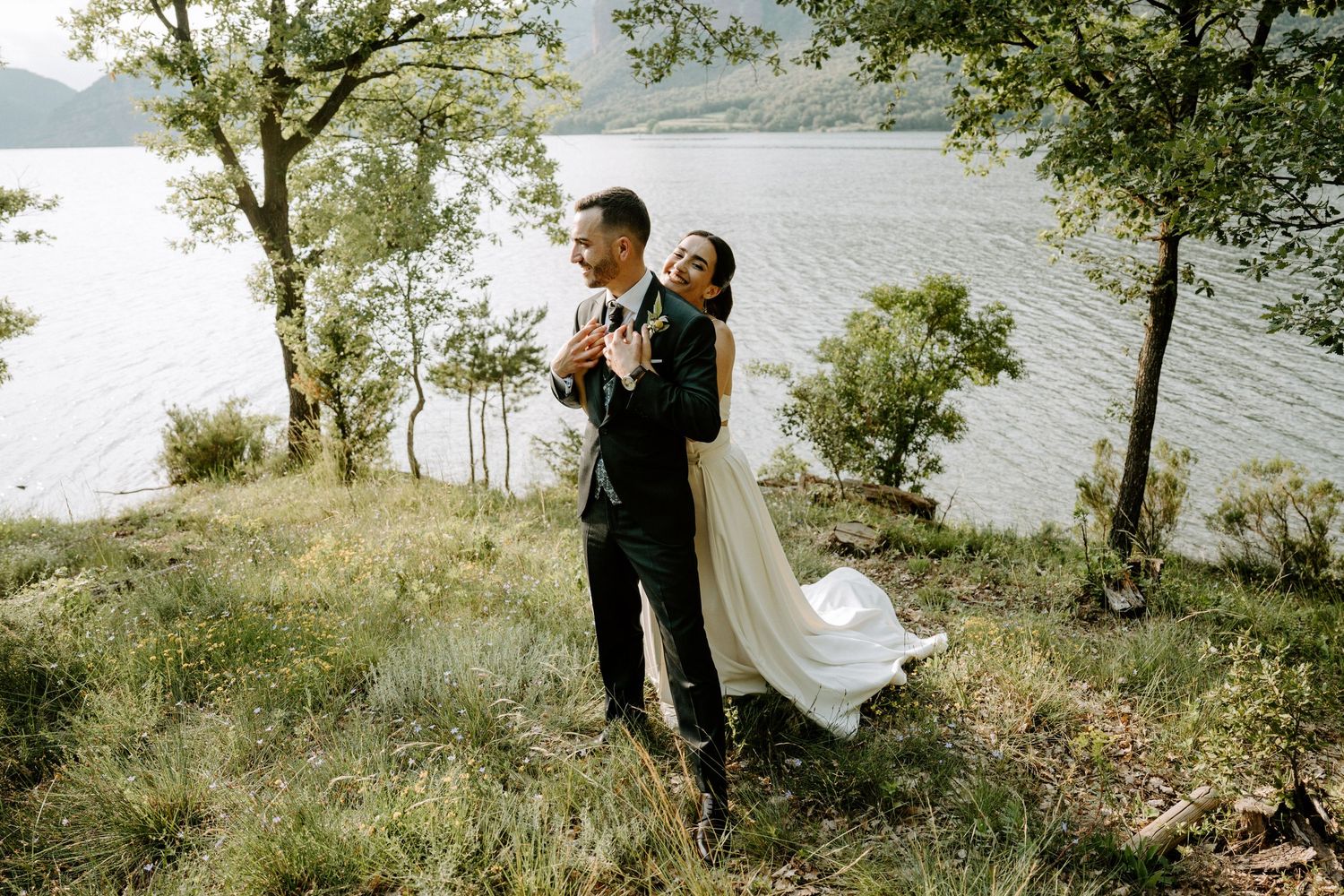 Romantic bride and groom embrace on scenic lakeside hillside surrounded by trees.