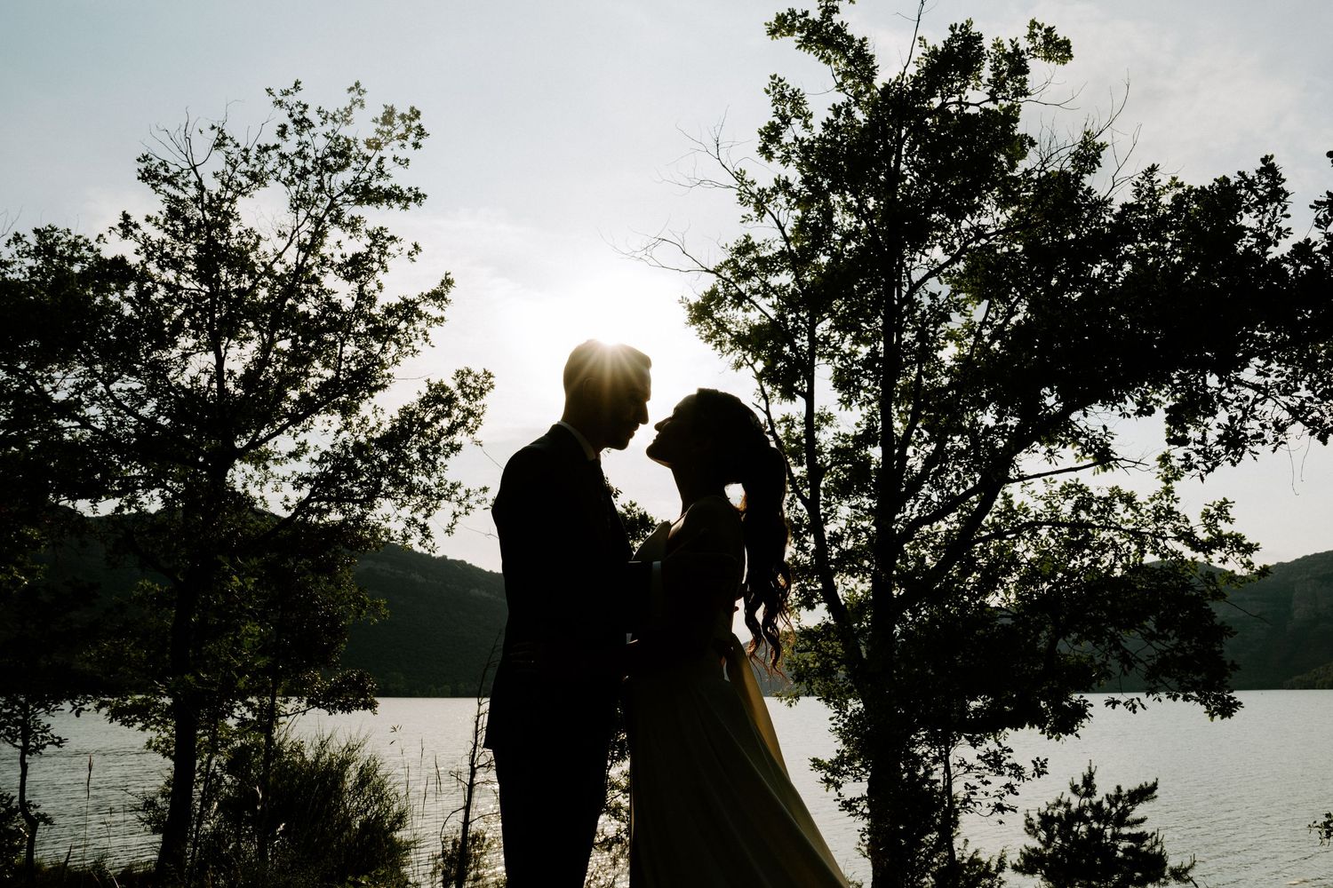 Romantic silhouette of bride and groom embracing near lake at sunset with trees framing scene.