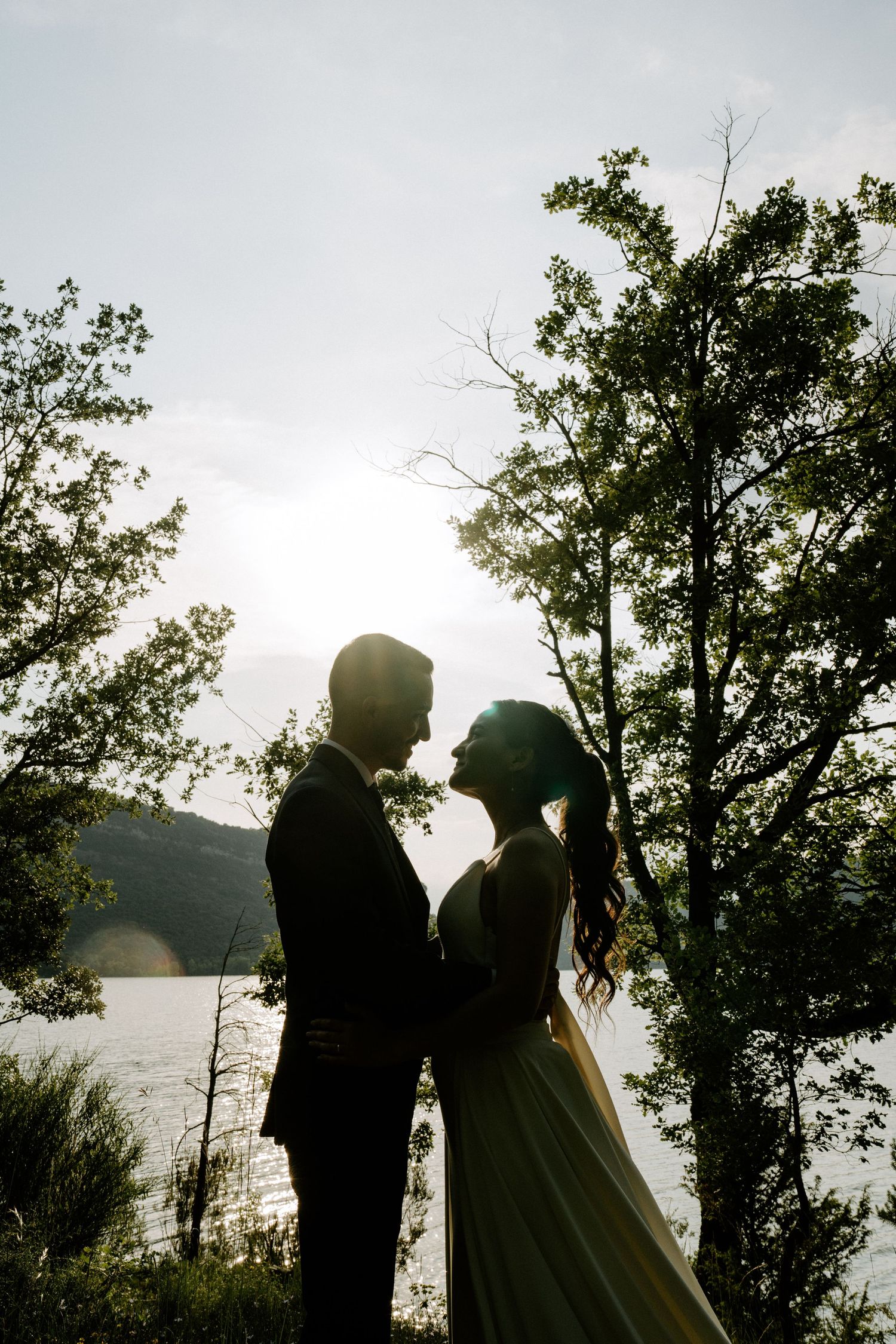 Romantic silhouette of bride and groom facing each other by a lake at sunset.