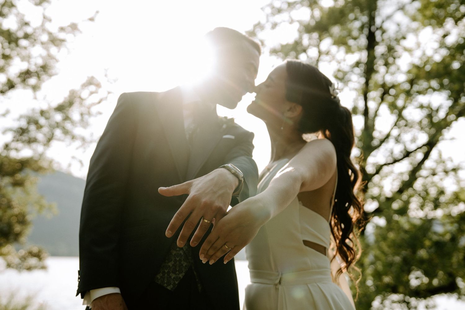 A bride and groom share a romantic kiss backlit by golden sunlight among trees at their wedding.