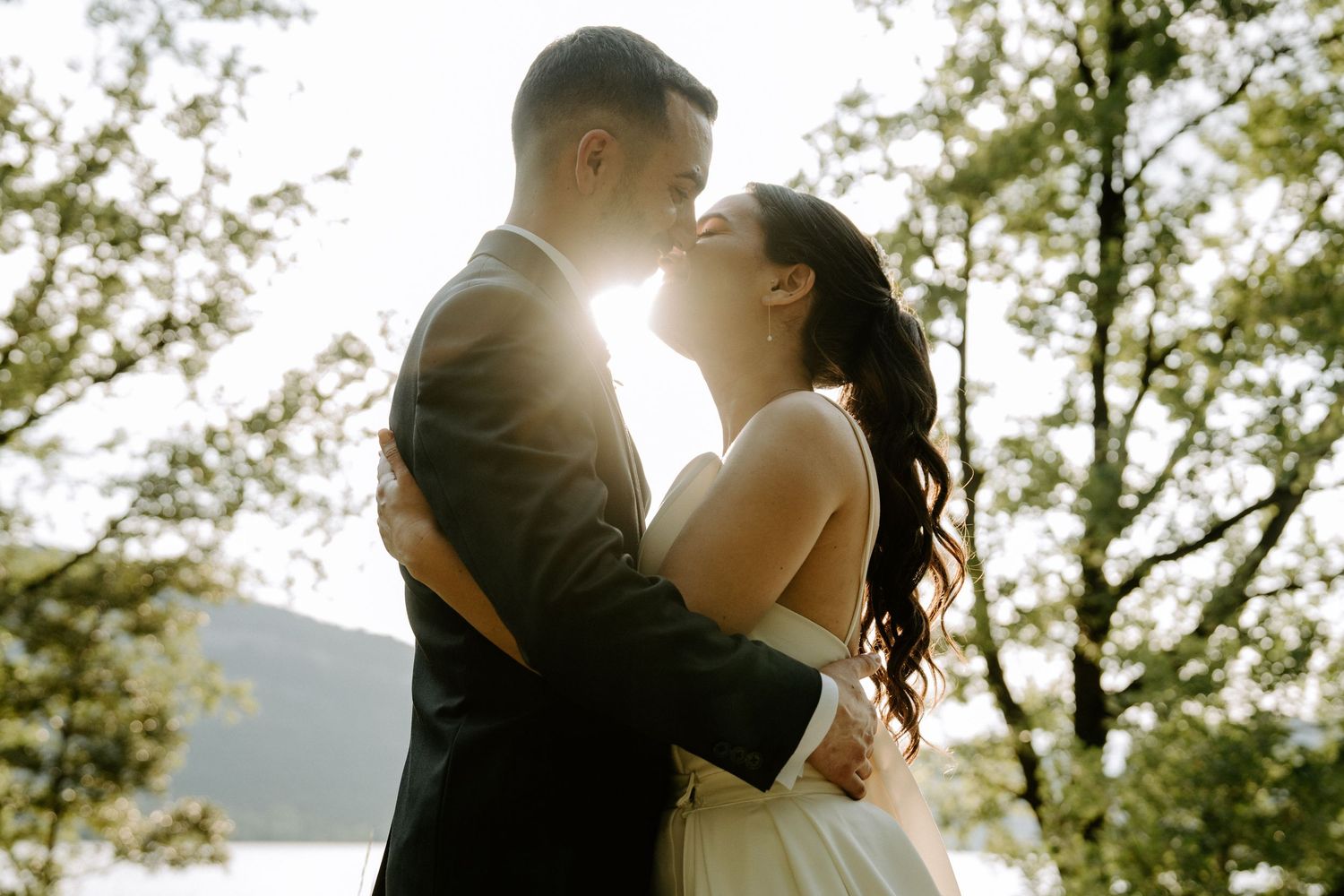 Bride and groom sharing a romantic kiss backlit by golden sunlight in a forest.