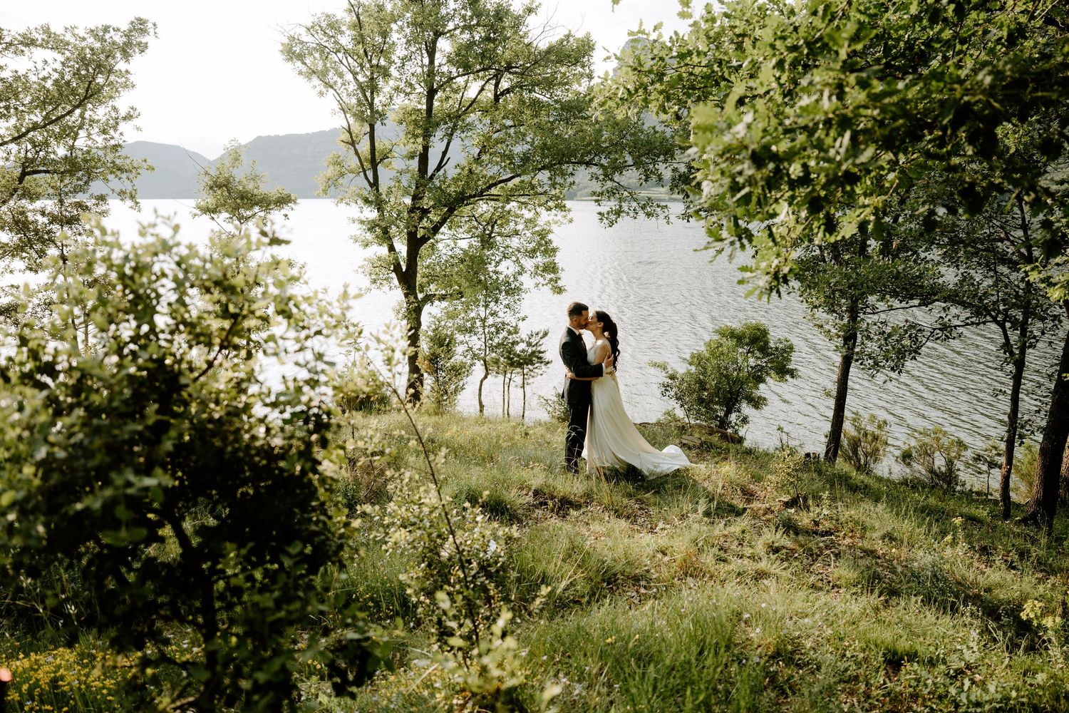Bride and groom embracing on grassy hillside overlooking scenic lake surrounded by lush green trees.