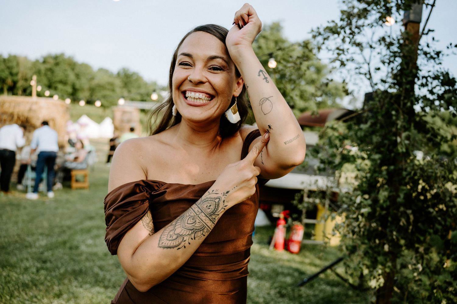 Smiling woman in brown off-shoulder dress showing arm tattoo at outdoor wedding reception.