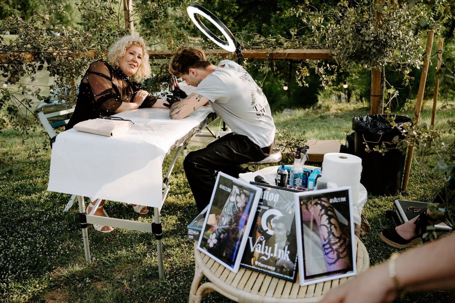 Wedding guest receives outdoor tattoo from artist at decorated table during garden reception.