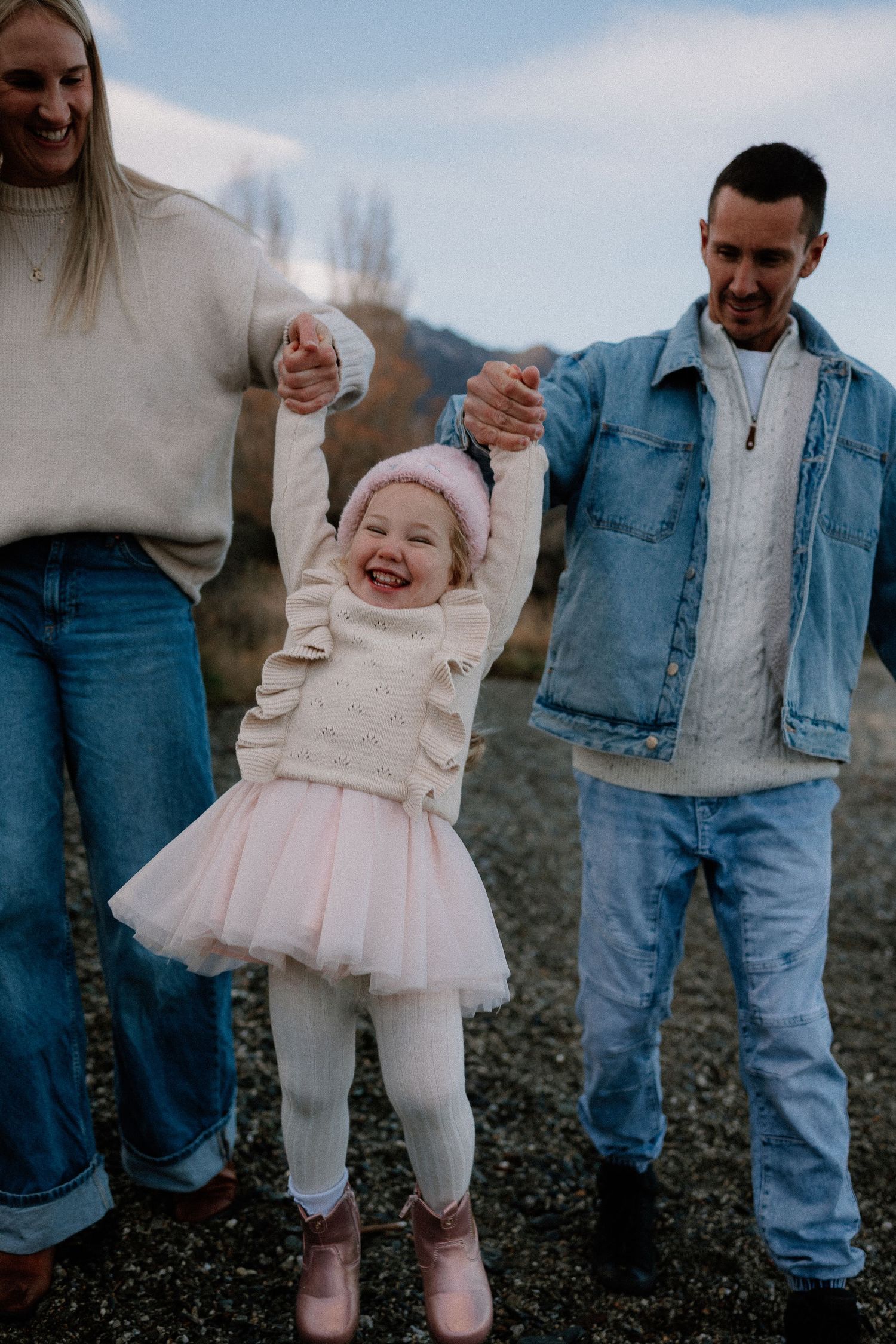 A joyful toddler girl in a pink tutu is lifted by her parents hands outdoors, laughing with delight.
