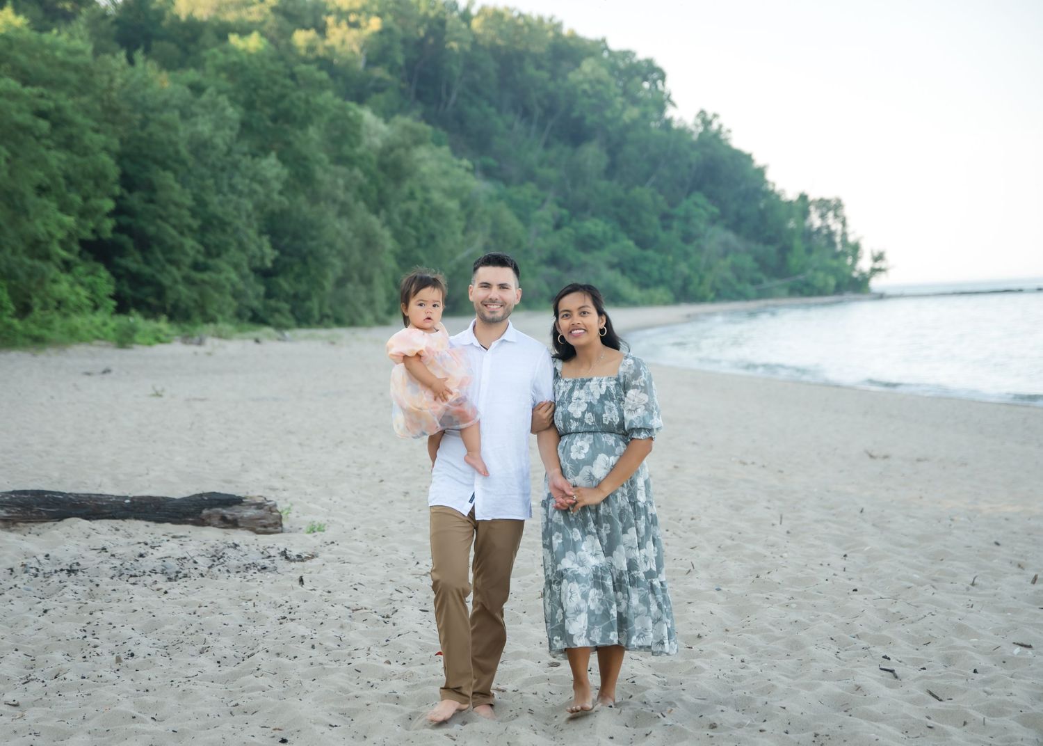 Family of three poses on a sandy beach, parents holding toddler, lush green trees and calm water in background.