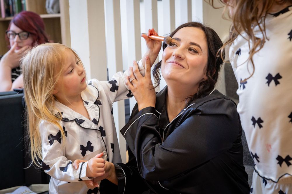 A bride smiles as a makeup artist applies cosmetics while a young girl in matching pajamas watches closely.
