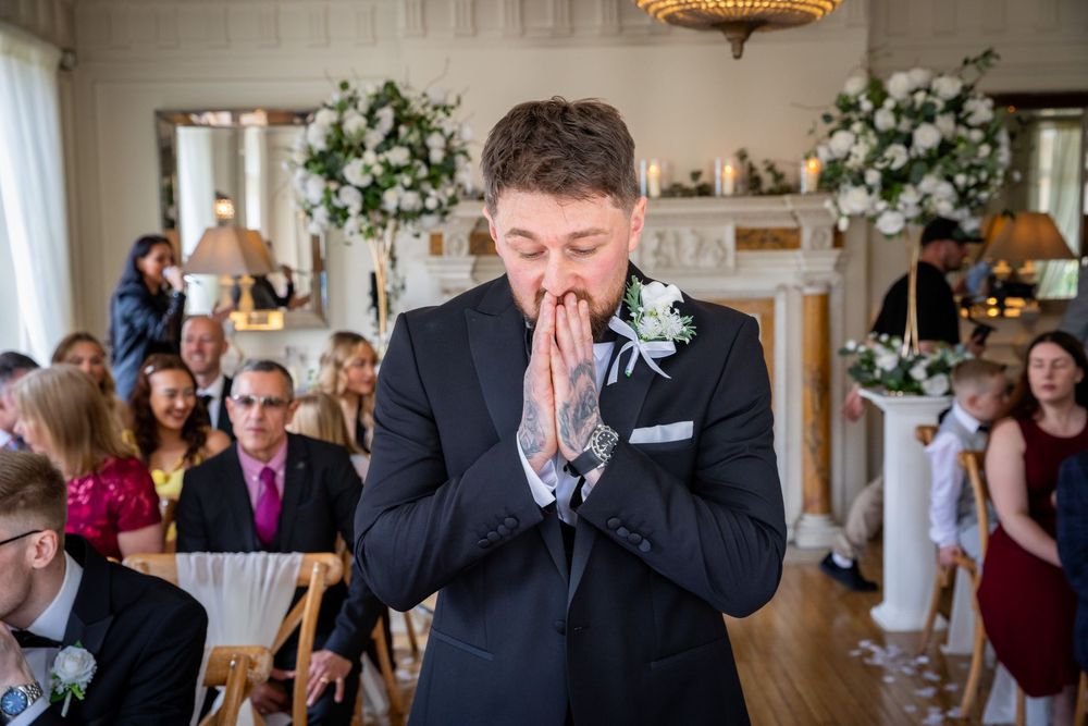 Emotional groom clasping hands in prayer during wedding ceremony, wearing black suit with white boutonniere.