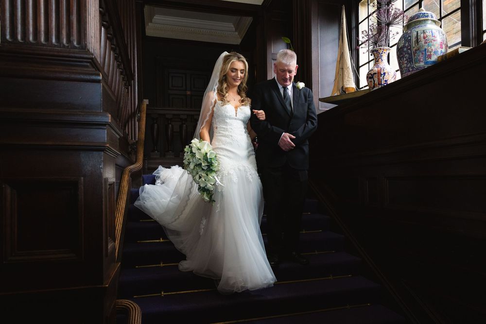 Bride in white gown descends grand wooden staircase with escort in suit, holding green bouquet in elegant venue.