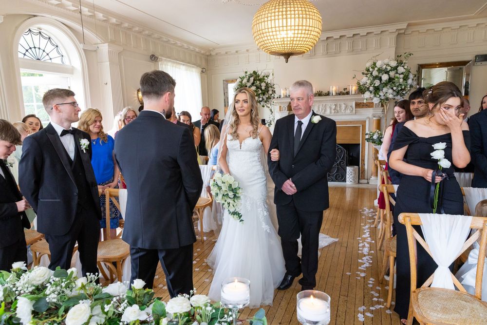 Bride walks down the aisle with her father in an elegant wedding venue decorated with white flowers and candles.