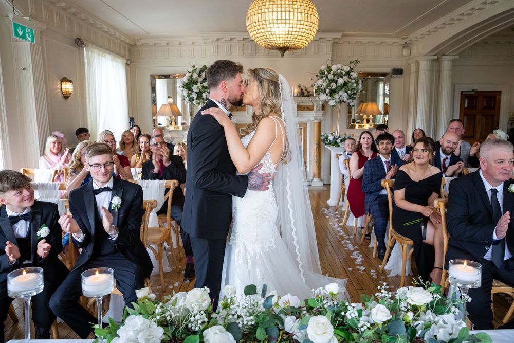 Bride and groom share first kiss at elegant wedding ceremony with guests applauding in decorated venue.